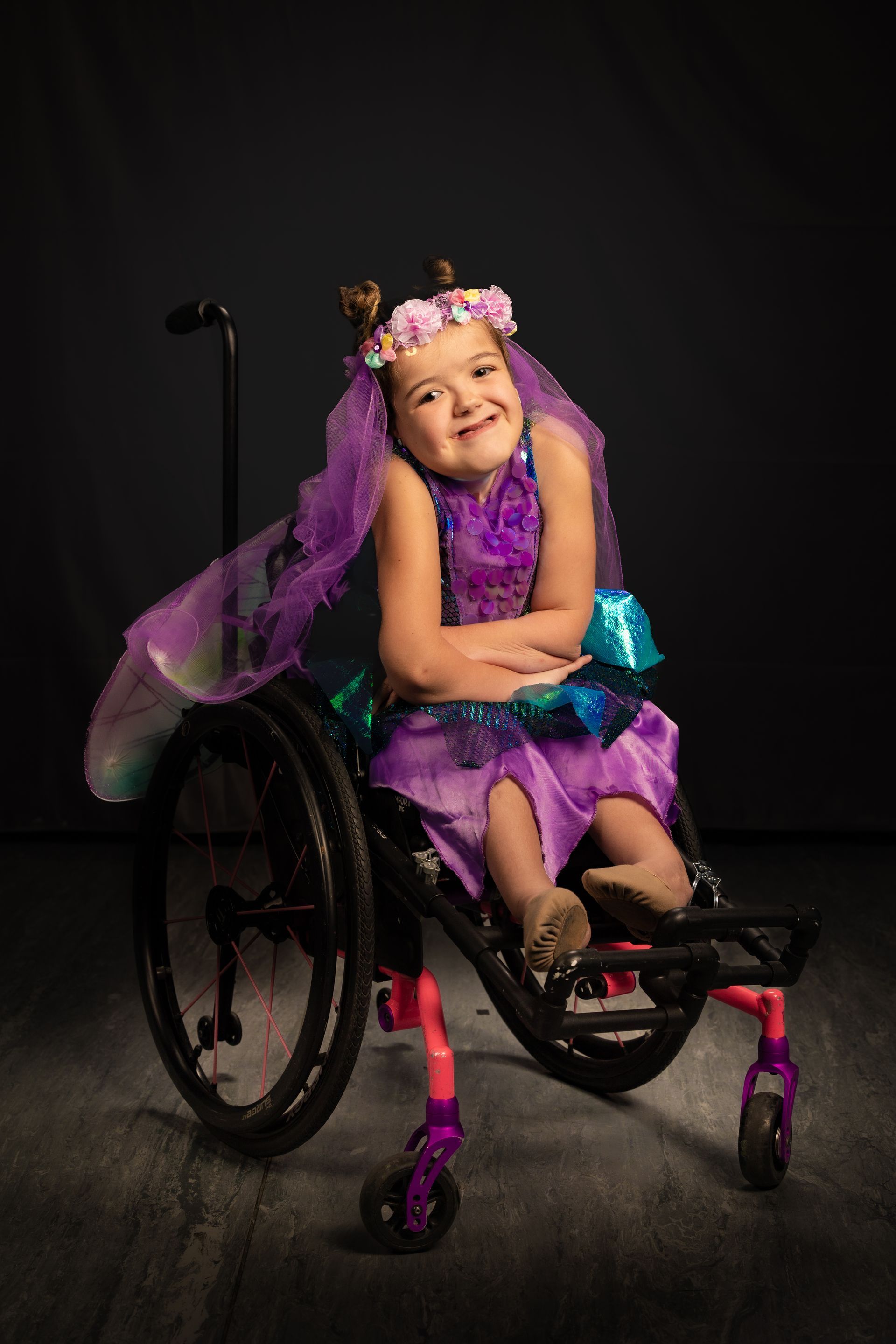 Child in a purple fairy costume seated in a wheelchair, smiling against a dark background.