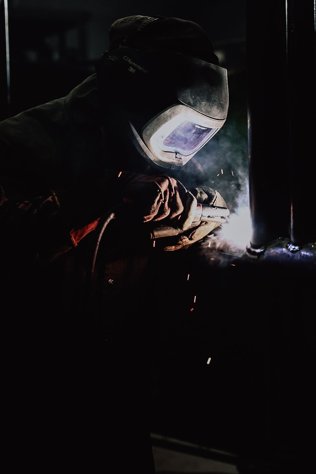 A welder wearing protective gear and a glowing helmet works on a metal project, creating sparks in a dark setting.