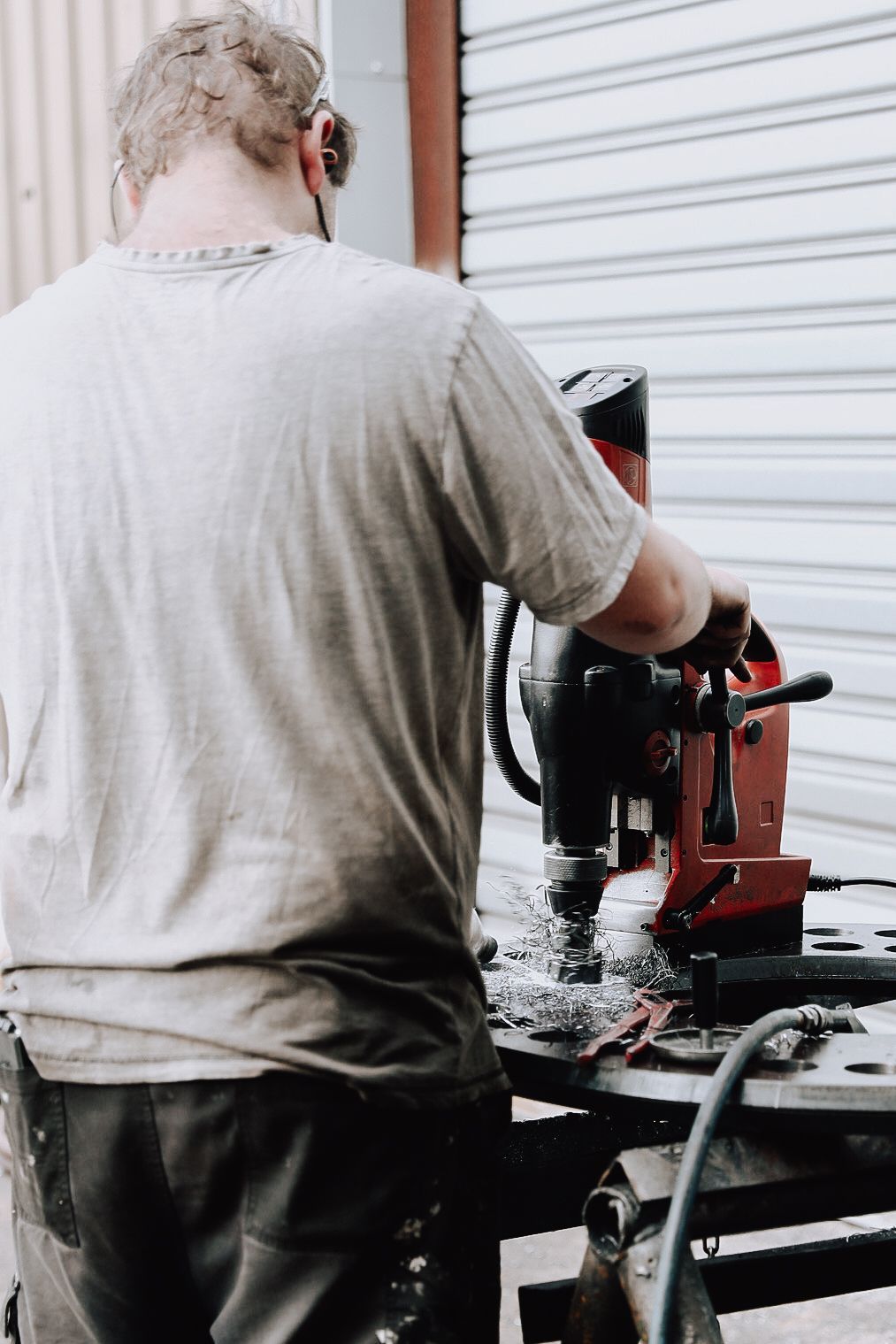 A person in a t-shirt operates a red magnetic drill press on a metal workbench in a workshop.