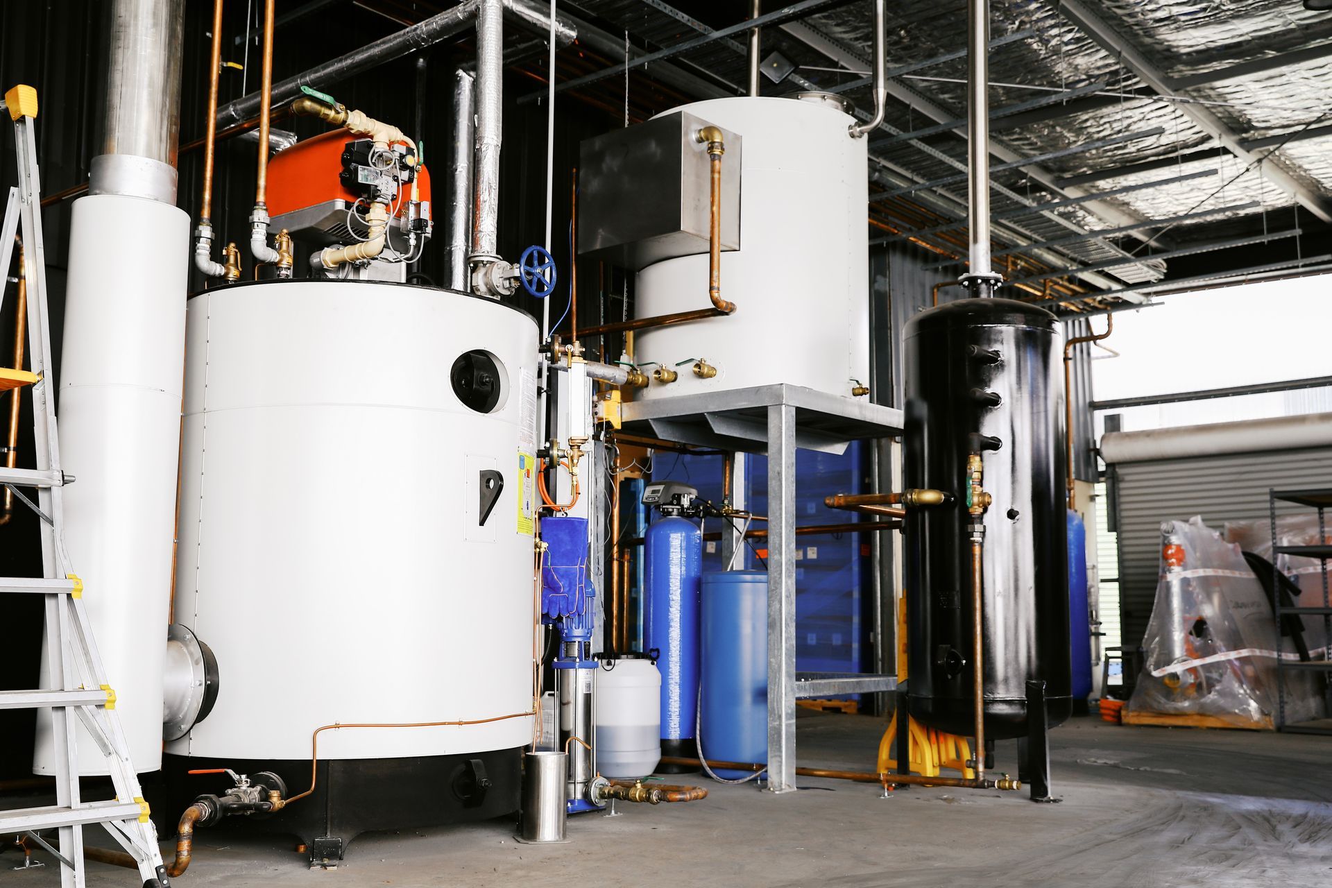 Industrial boiler system with white and black tanks, pipes, and control equipment in a workshop setting.