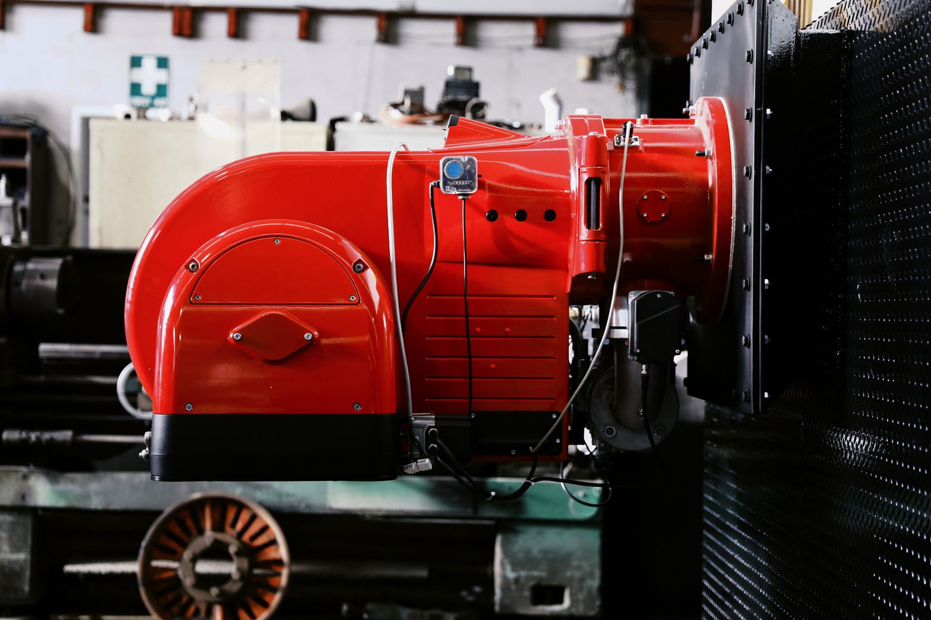 A bright red industrial burner unit mounted on a black metal plate in a workshop setting.