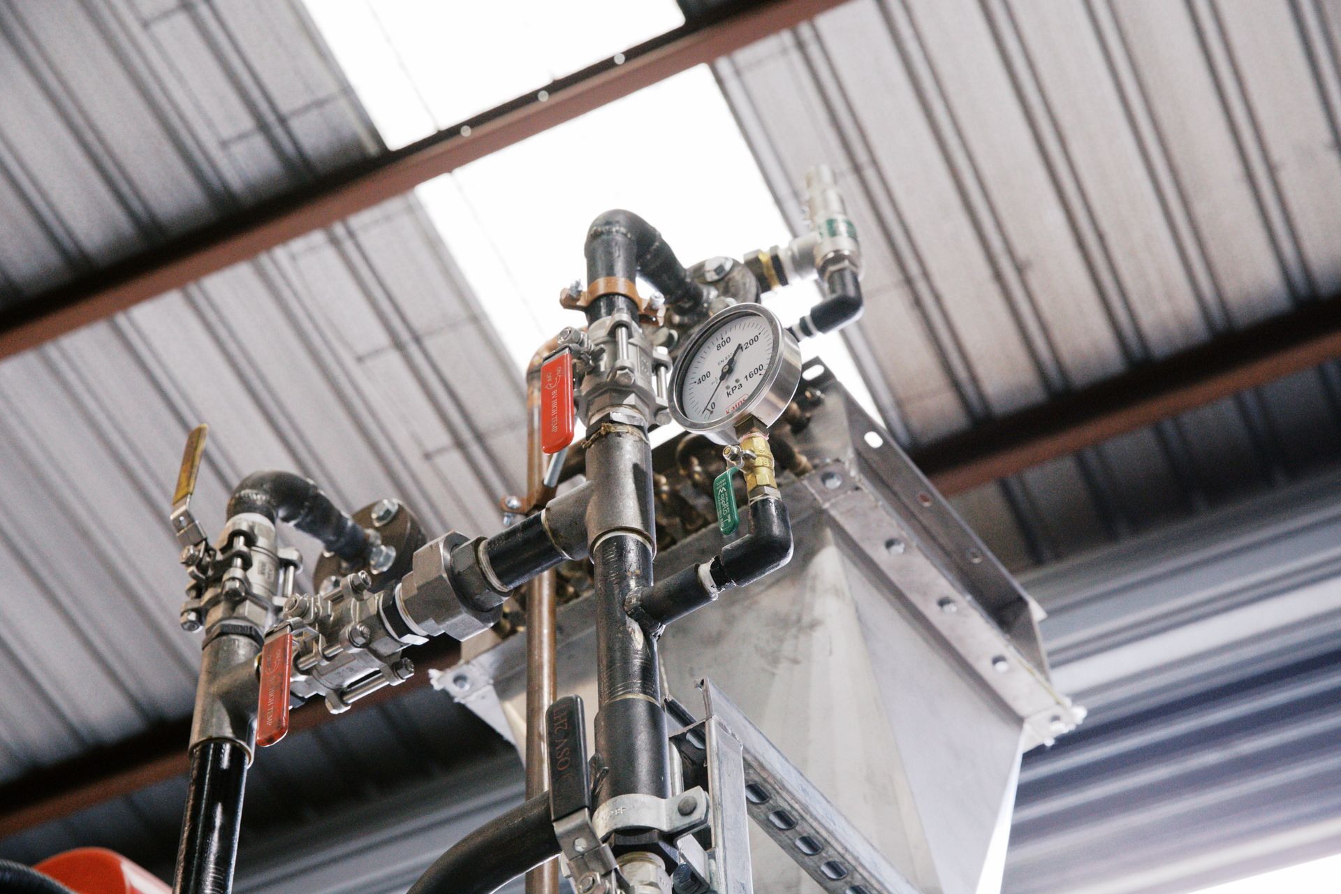 A close-up of industrial machinery featuring black pipes, red-handled valves, and a circular pressure gauge under a roof. A close-up of industrial machinery featuring black pipes, red-handled valves, and a circular pressure gauge under a roof.