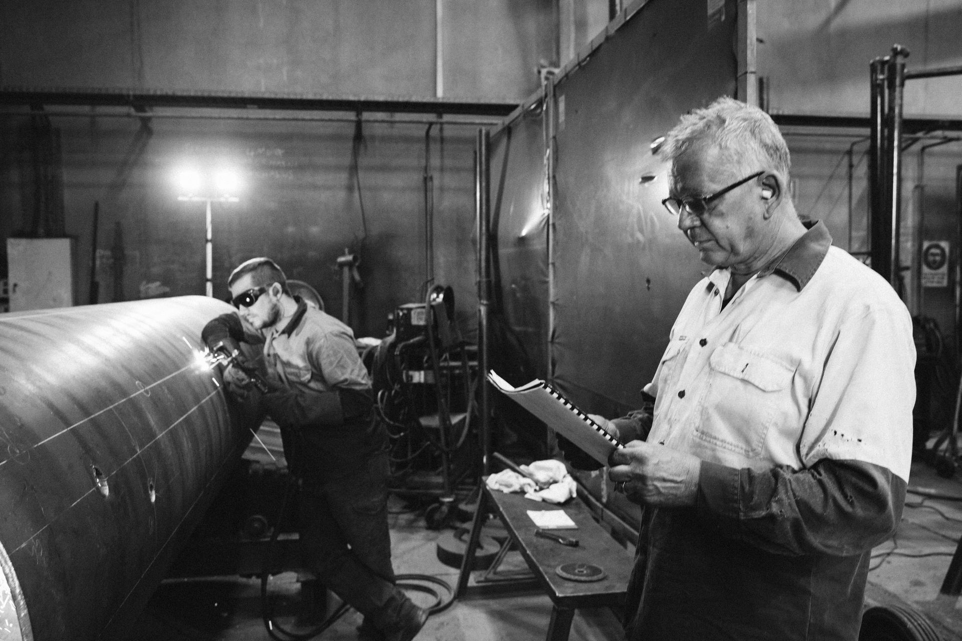 An industrial worker welds a large pipe while a colleague holds a document and monitors the process in a workshop. An industrial worker welds a large pipe while a colleague holds a document and monitors the process in a workshop.