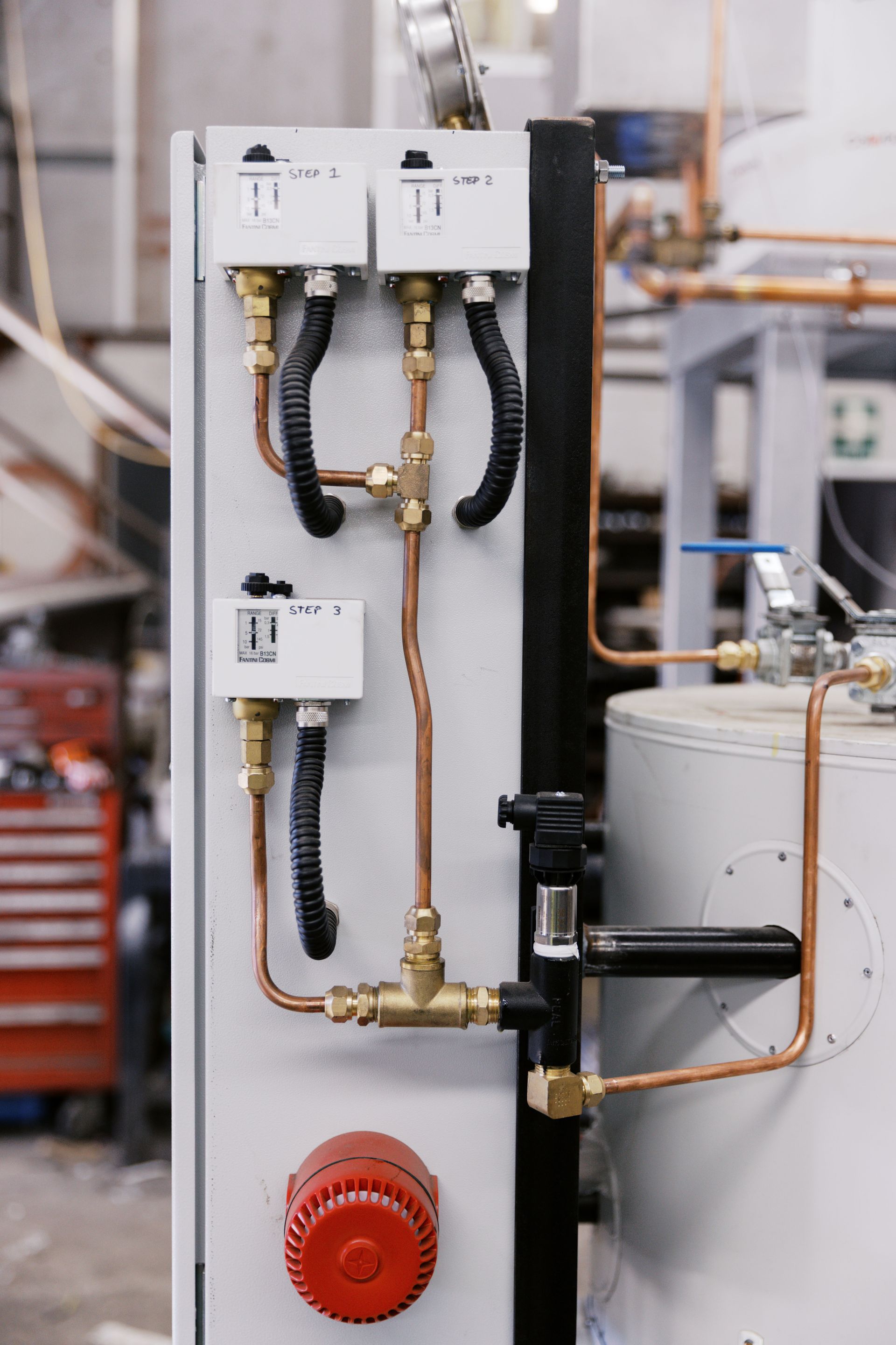 A control panel with three electronic sensors, copper tubing, and a red circular alarm mounted on a white industrial unit.