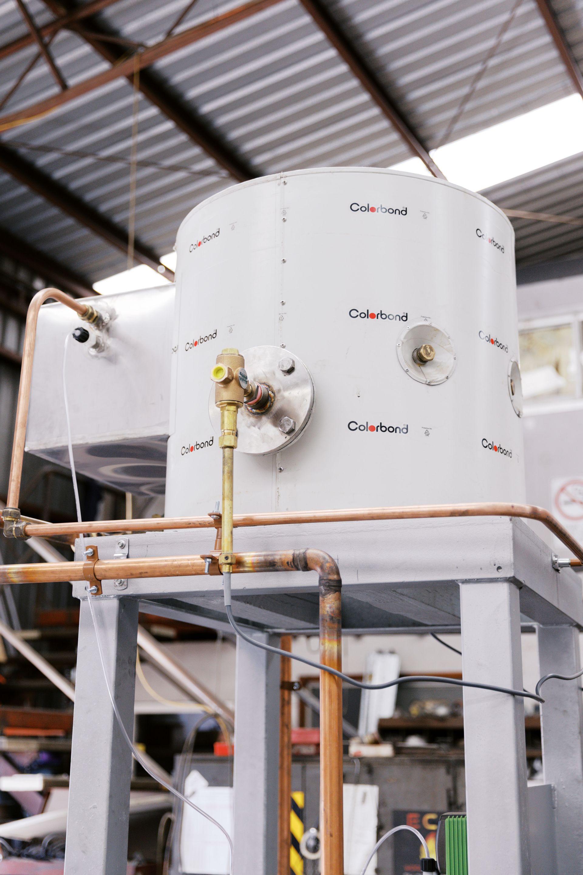 A large, cylindrical industrial metal tank mounted on a grey frame, featuring copper piping and valves in a workshop.