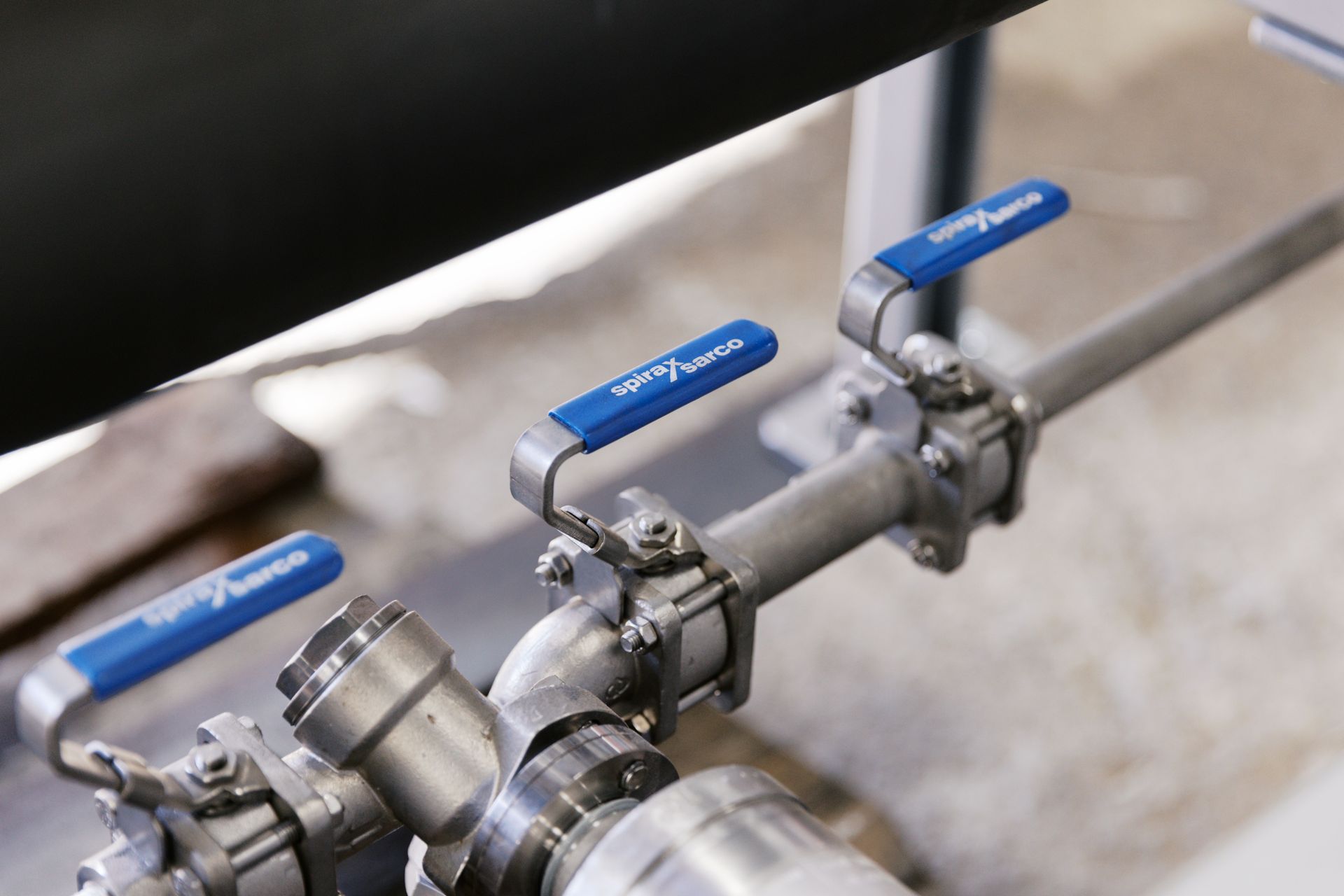 Three stainless steel pipes with blue-handled ball valves mounted on a metal frame. Three stainless steel pipes with blue-handled ball valves mounted on a metal frame.