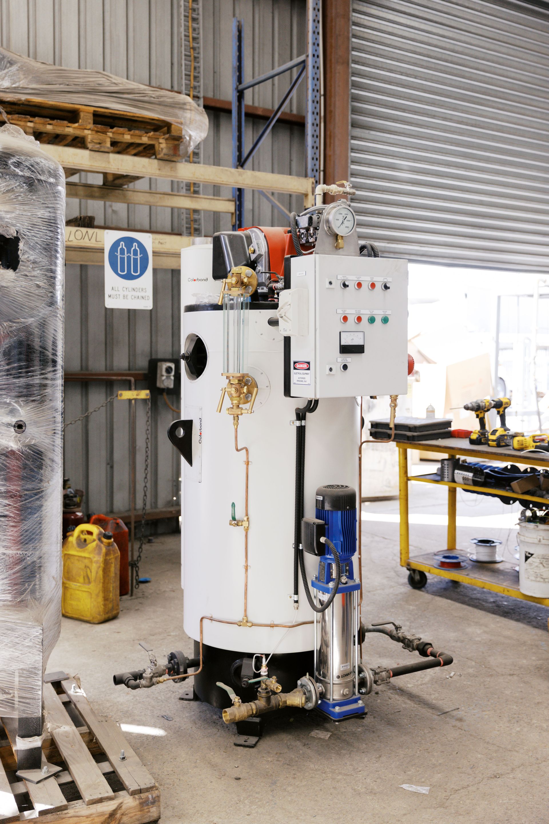 An industrial white cylindrical boiler with a blue pump and control box stands in a workshop setting.
