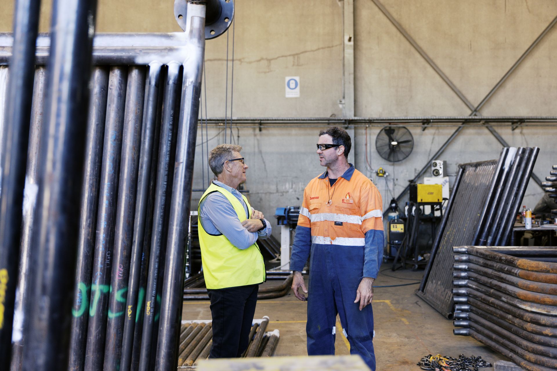 Two people in high-visibility work gear talk in an industrial warehouse filled with metal pipe components. Two people in high-visibility work gear talk in an industrial warehouse filled with metal pipe components.