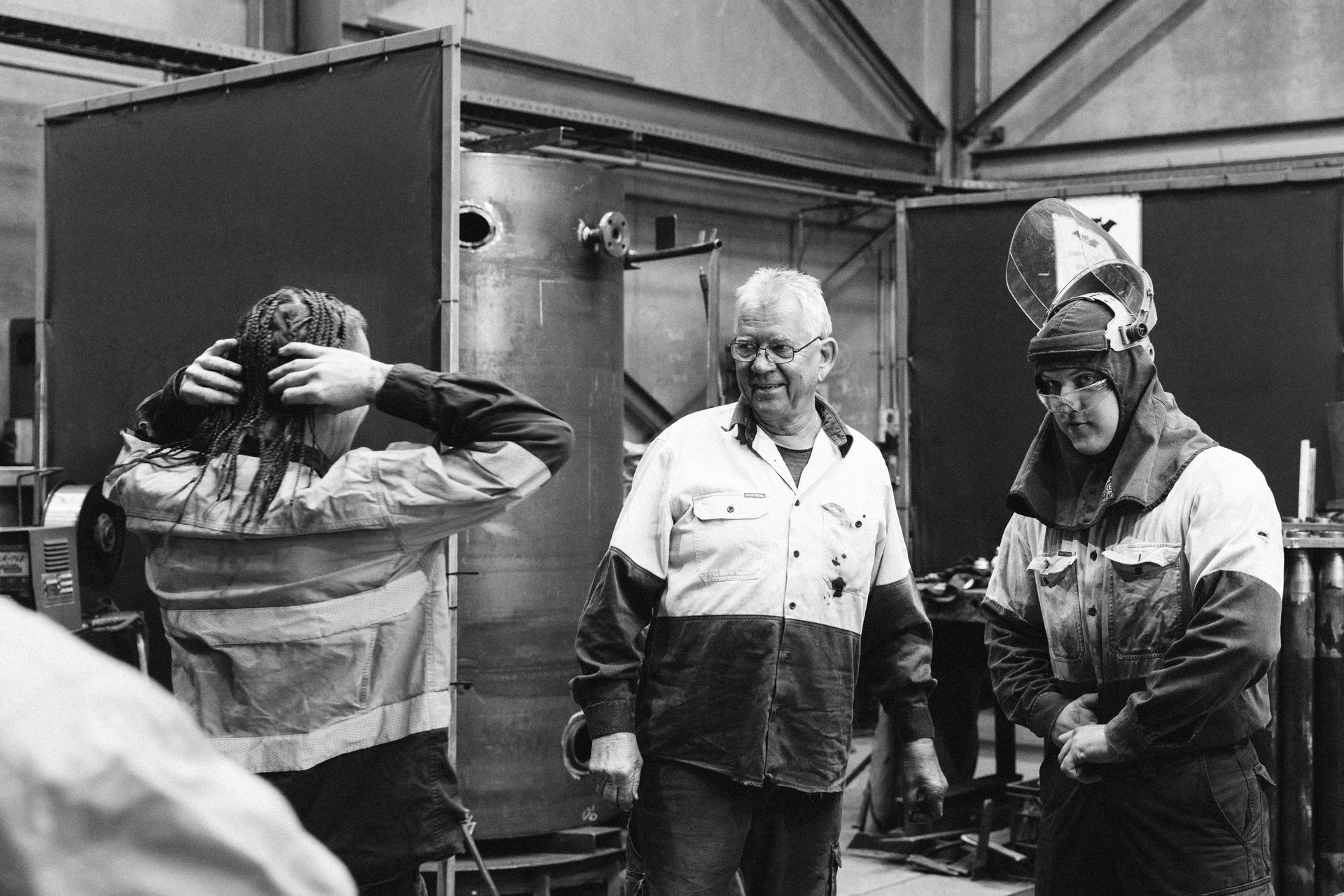 Three people in work uniforms standing in an industrial workshop; one person adjusts their hair, two others converse. Three people in work uniforms standing in an industrial workshop; one person adjusts their hair, two others converse.