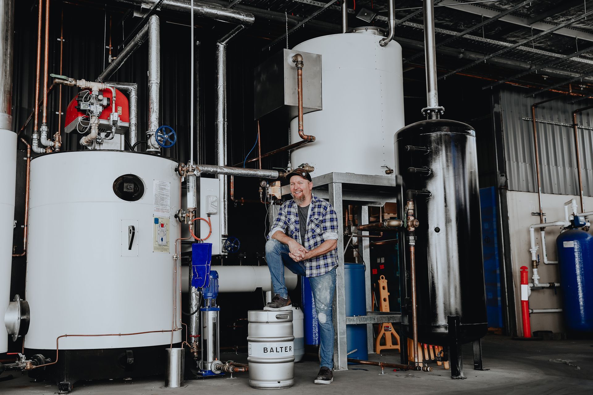 A person in a plaid shirt stands in an industrial facility, resting a foot on a metal keg between two large tanks.
