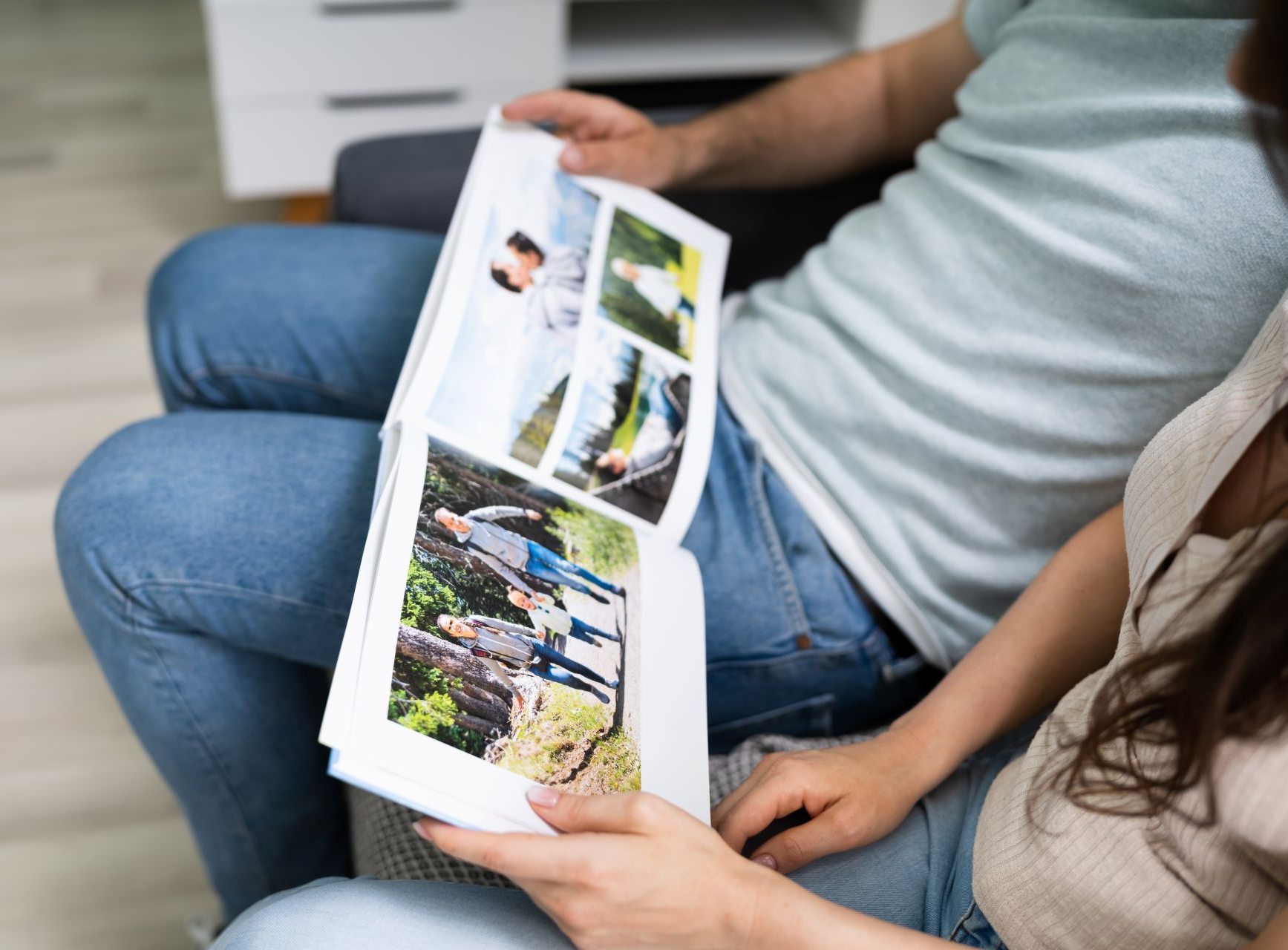 A person in a light blue t-shirt and jeans sits with another, holding open a photo book featuring pictures of a family.