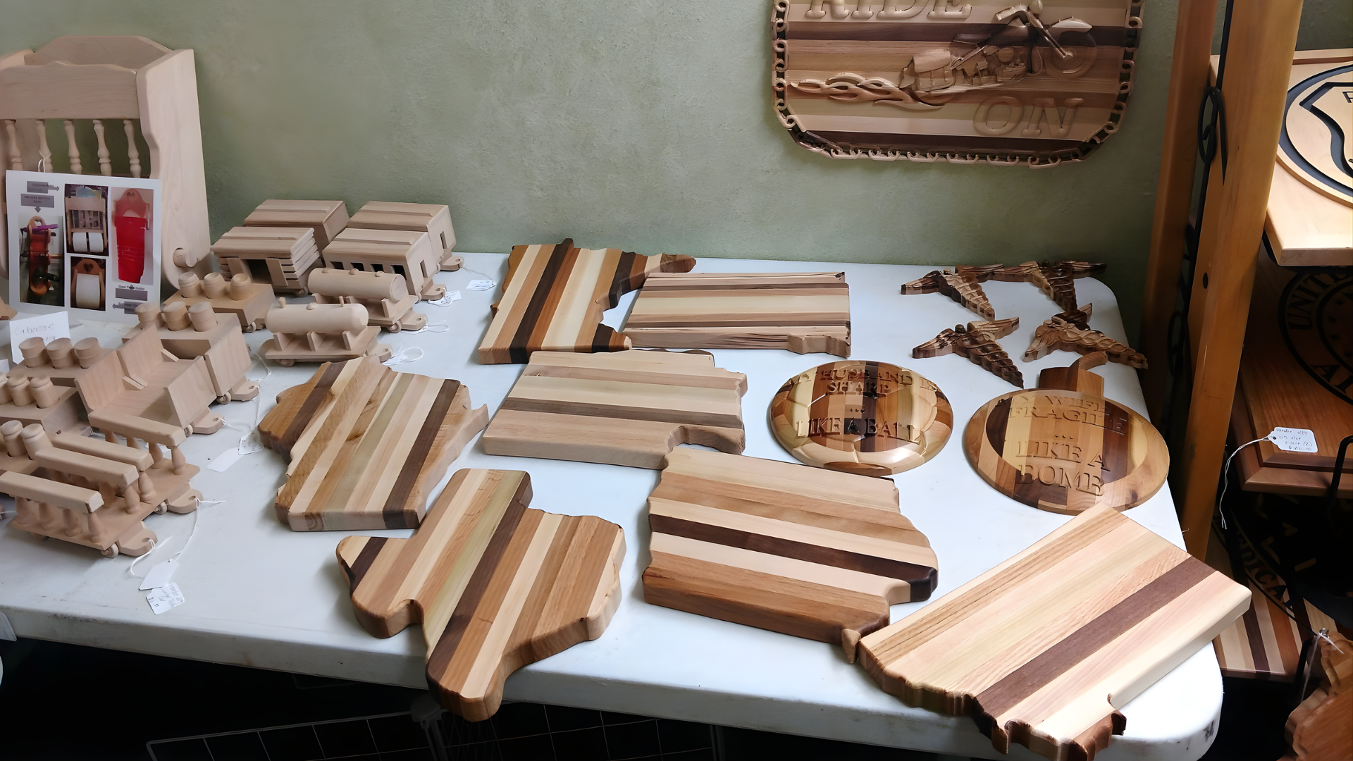 A white table displays handmade wooden crafts, including state-shaped cutting boards, small train cars, and round trays.