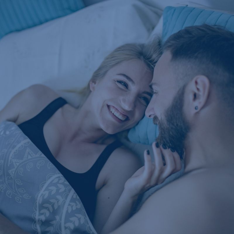 Blue-tinted overhead view of a smiling couple looking at each other while lying in bed.