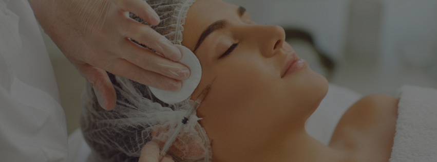 Close-up of a woman with her eyes closed receiving a cosmetic facial injection from a practitioner holding a cotton pad.