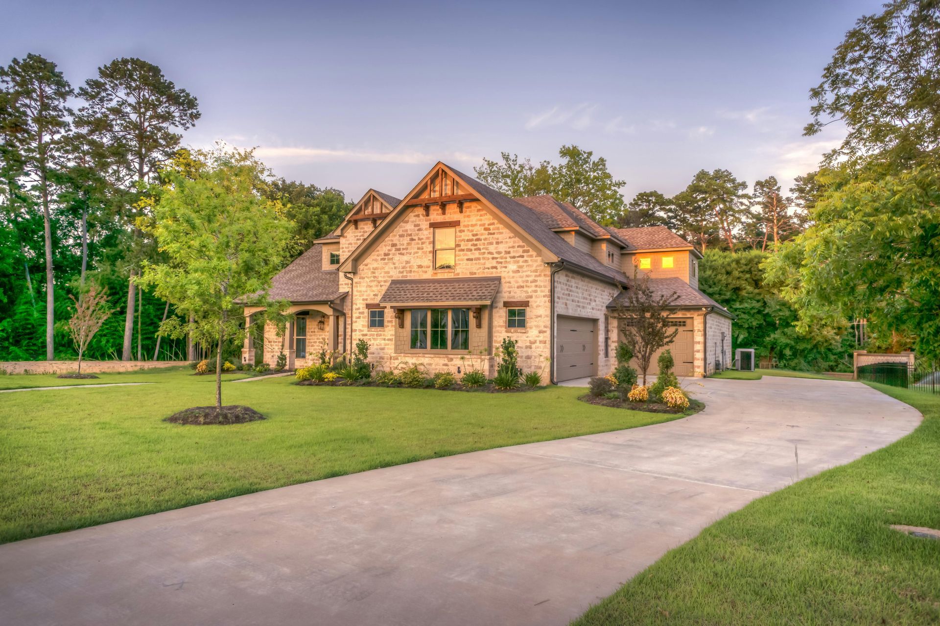Stone-brick house with a winding concrete driveway, green lawn, and surrounding trees under a blue sky.