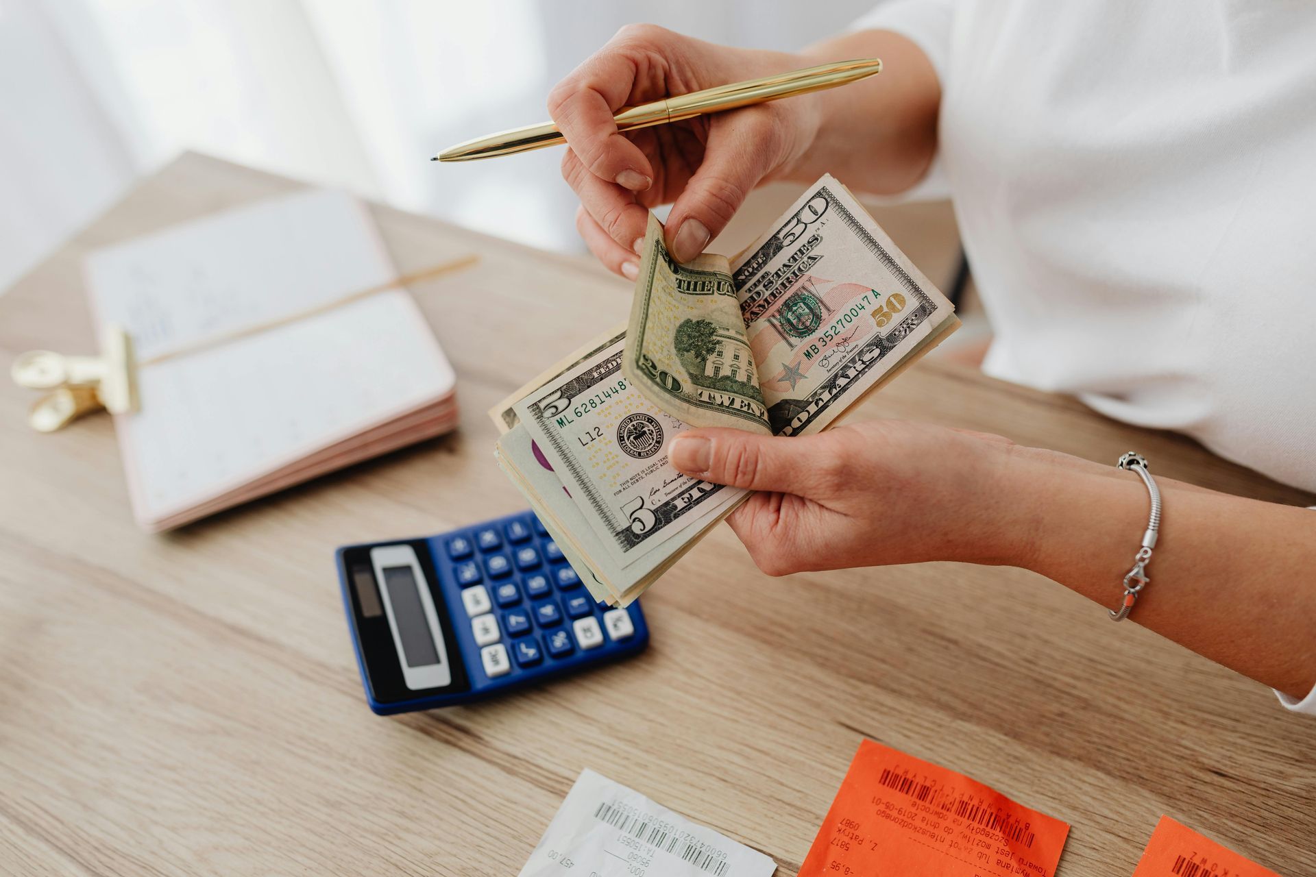 Hands counting U.S. dollar bills at a desk, with a calculator and notepad nearby.