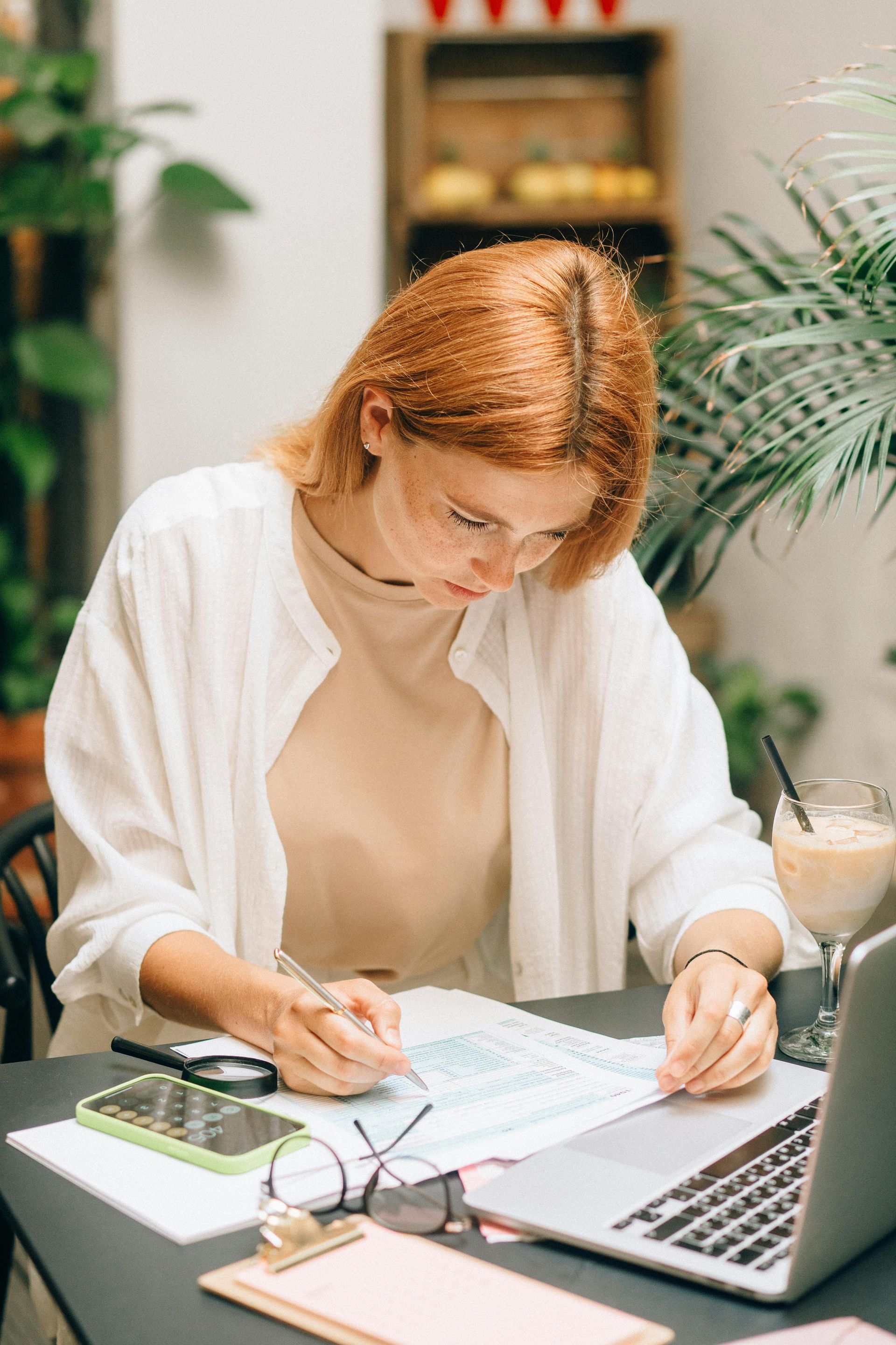 Woman with red hair and glasses working at a table, using a laptop and documents outdoors.