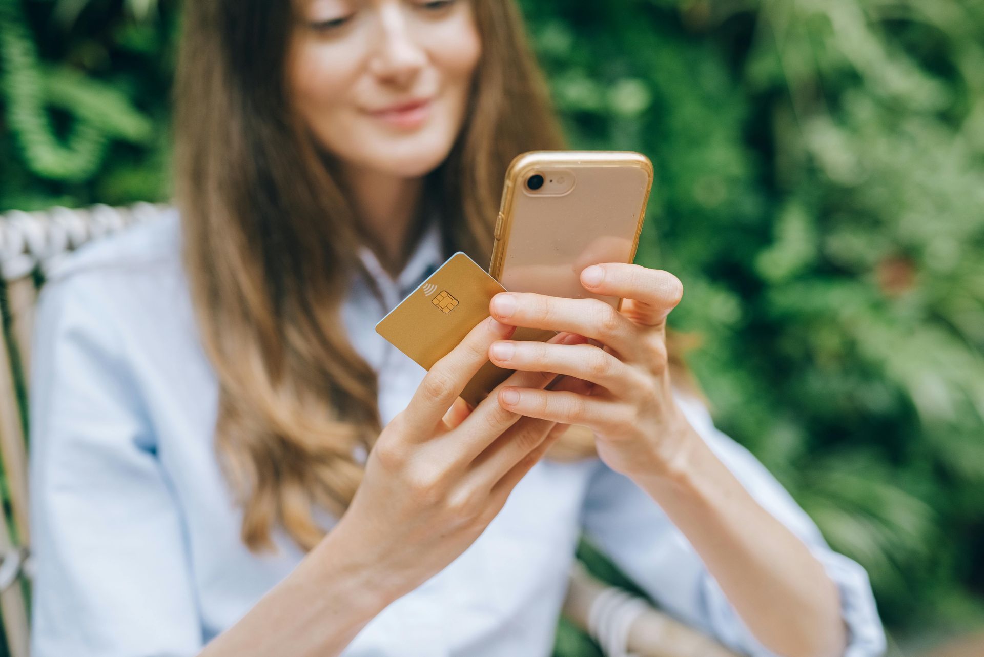 Woman holding a gold credit card and smartphone, smiling, outdoors with greenery in background.