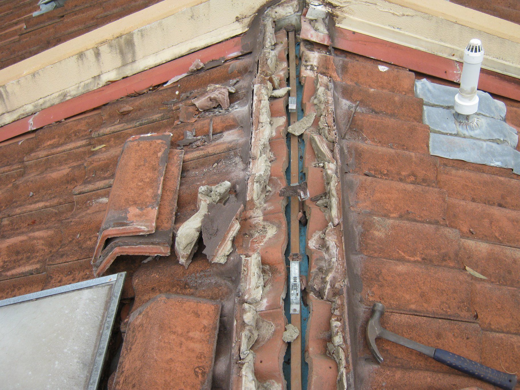 Damaged Roof With Missing Tiles and Deteriorated Sealant in a Valley. Hammer Rests Nearby.