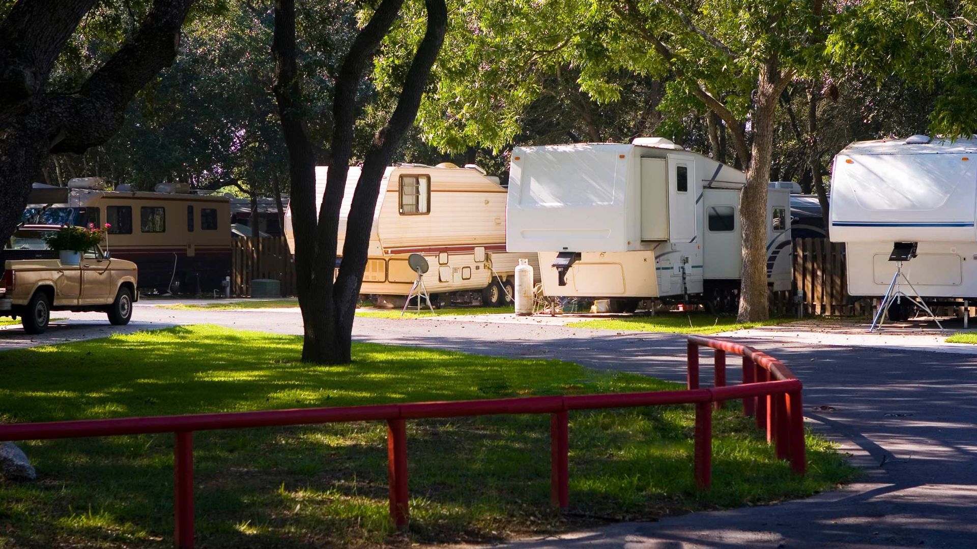 RVs parked in a shady campground with trees, a red railing, and a pickup truck in the background