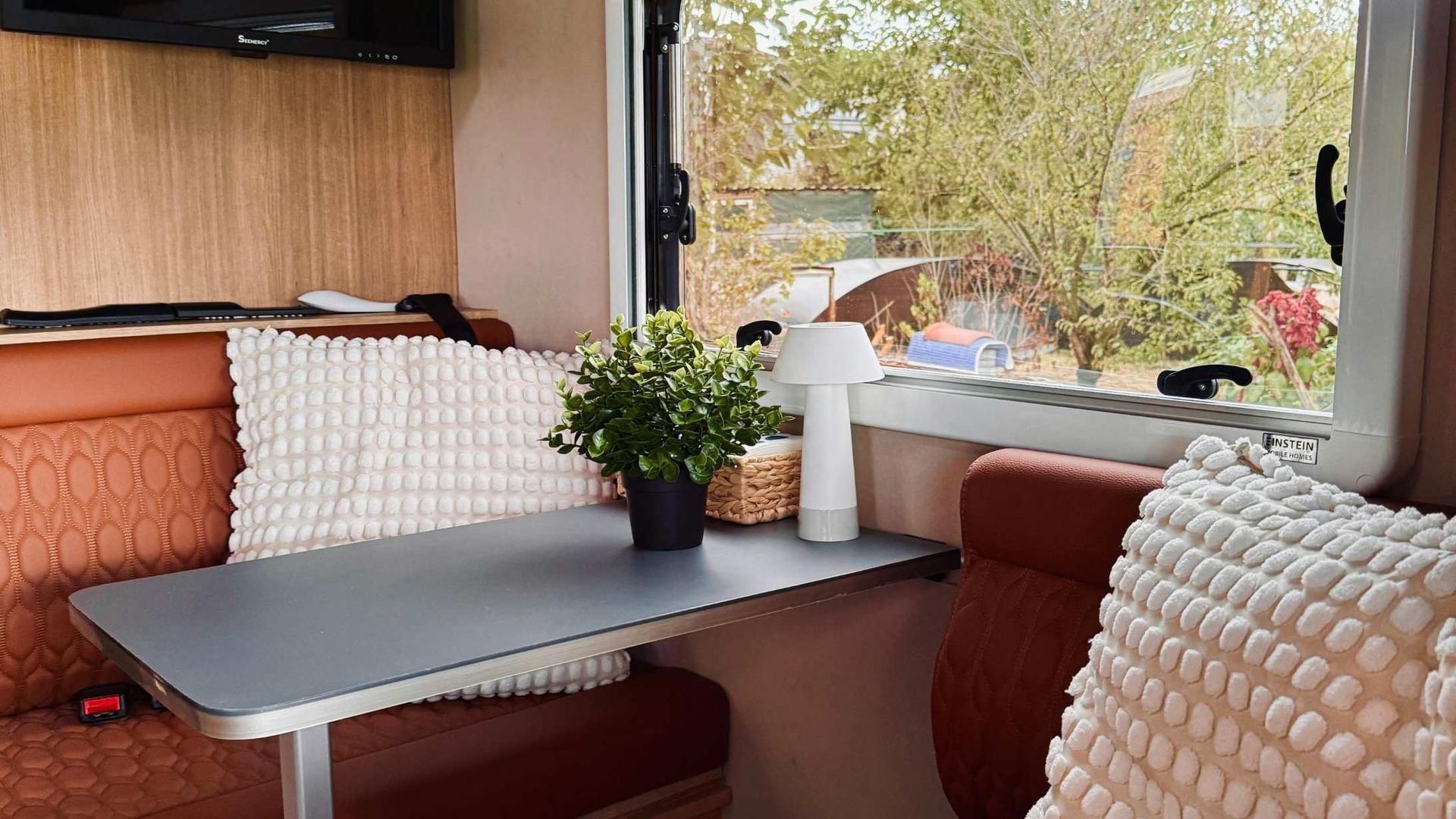 Cozy camper dining nook with a gray table, brown seats, white cushions, and a window view of trees