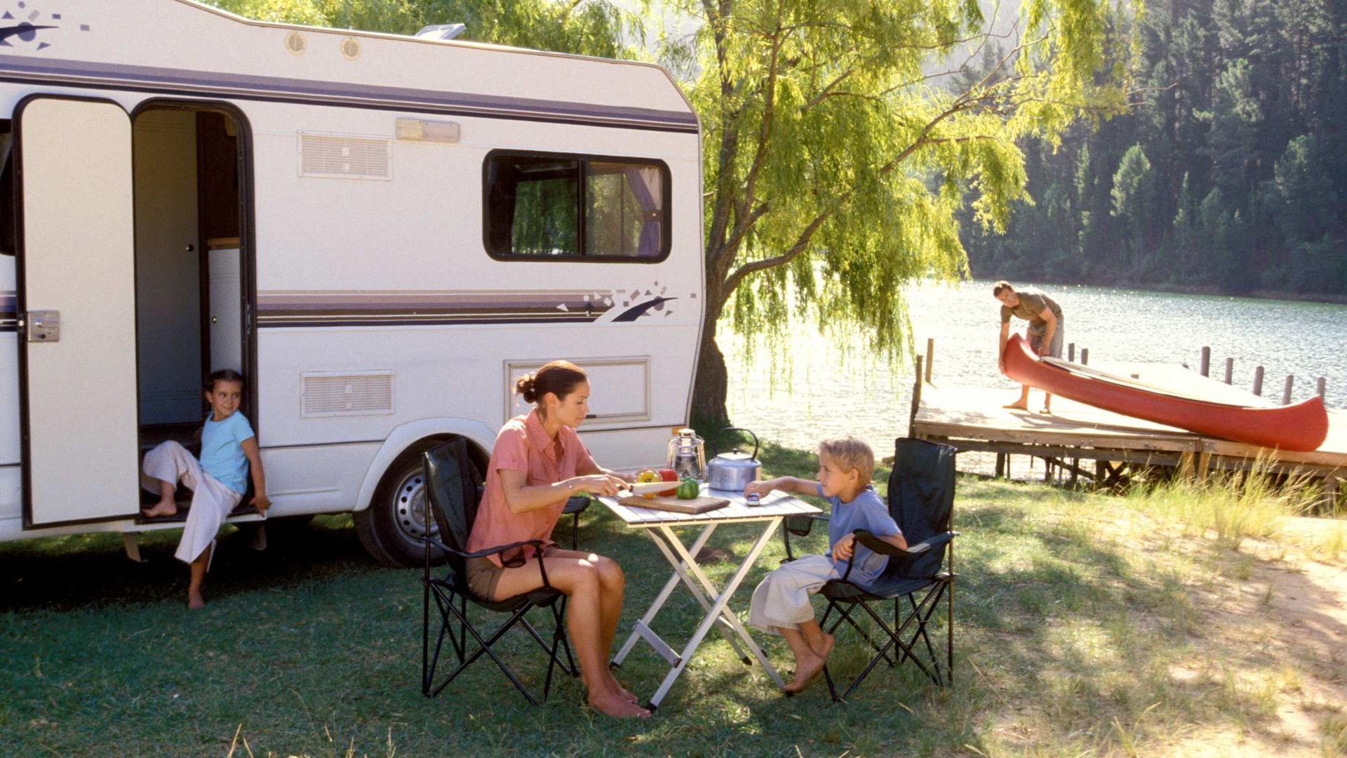 Family sitting outside a camper by a lakeside, with a canoe on the shore and trees in the background