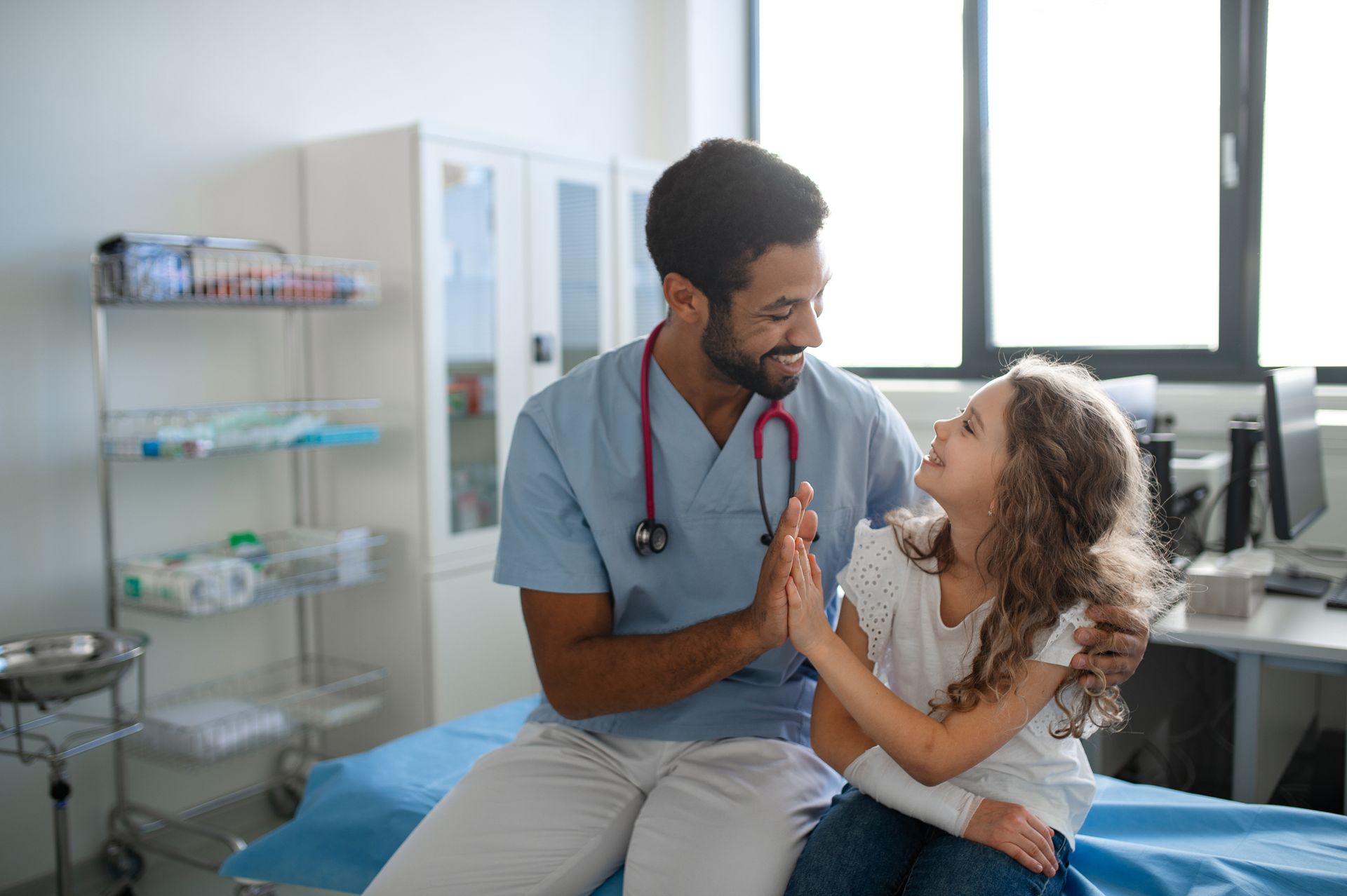 A doctor is sitting on a bed with a little girl and giving her a high five.