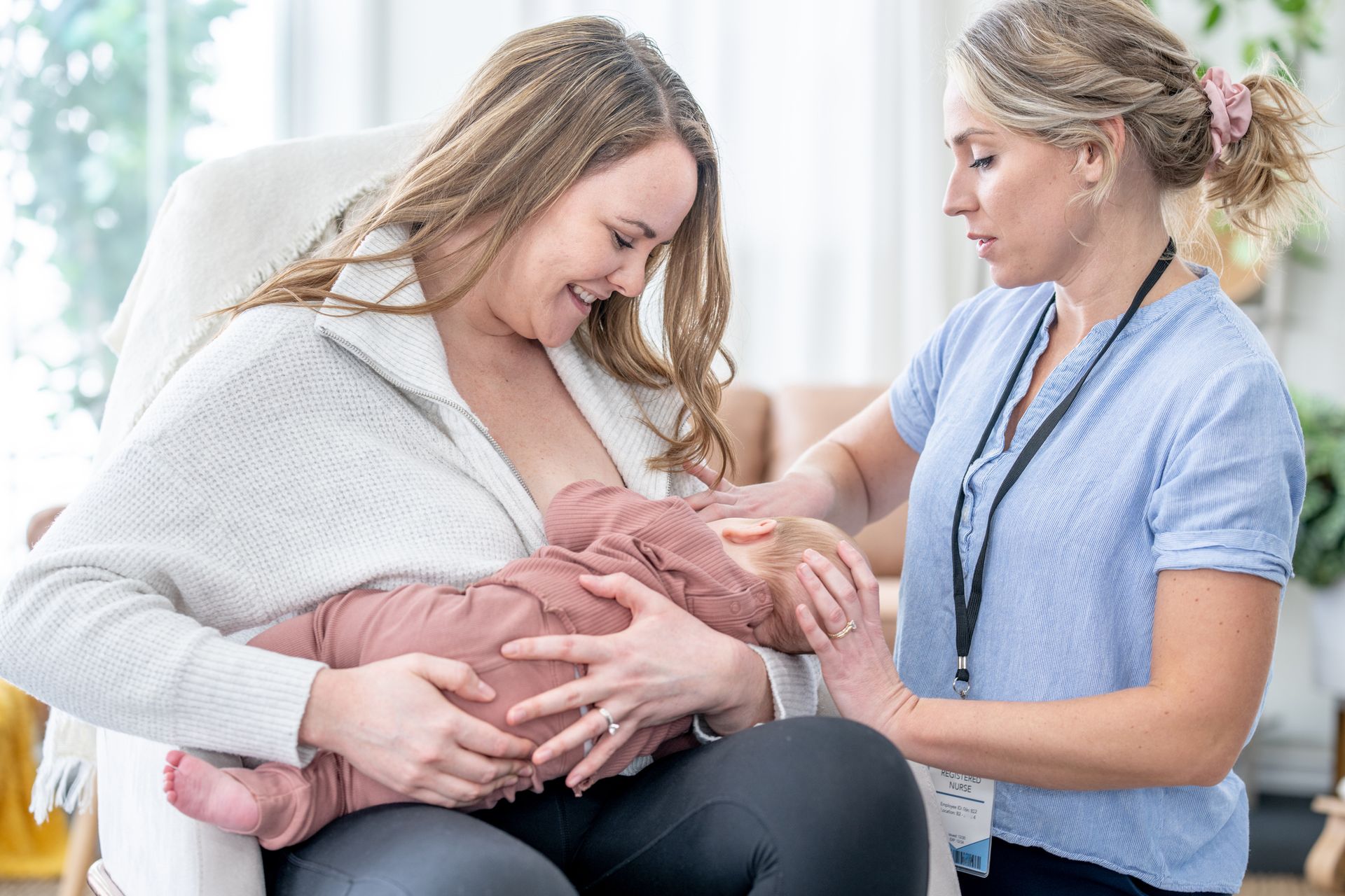 A woman is breastfeeding her baby in a chair while a nurse looks on.