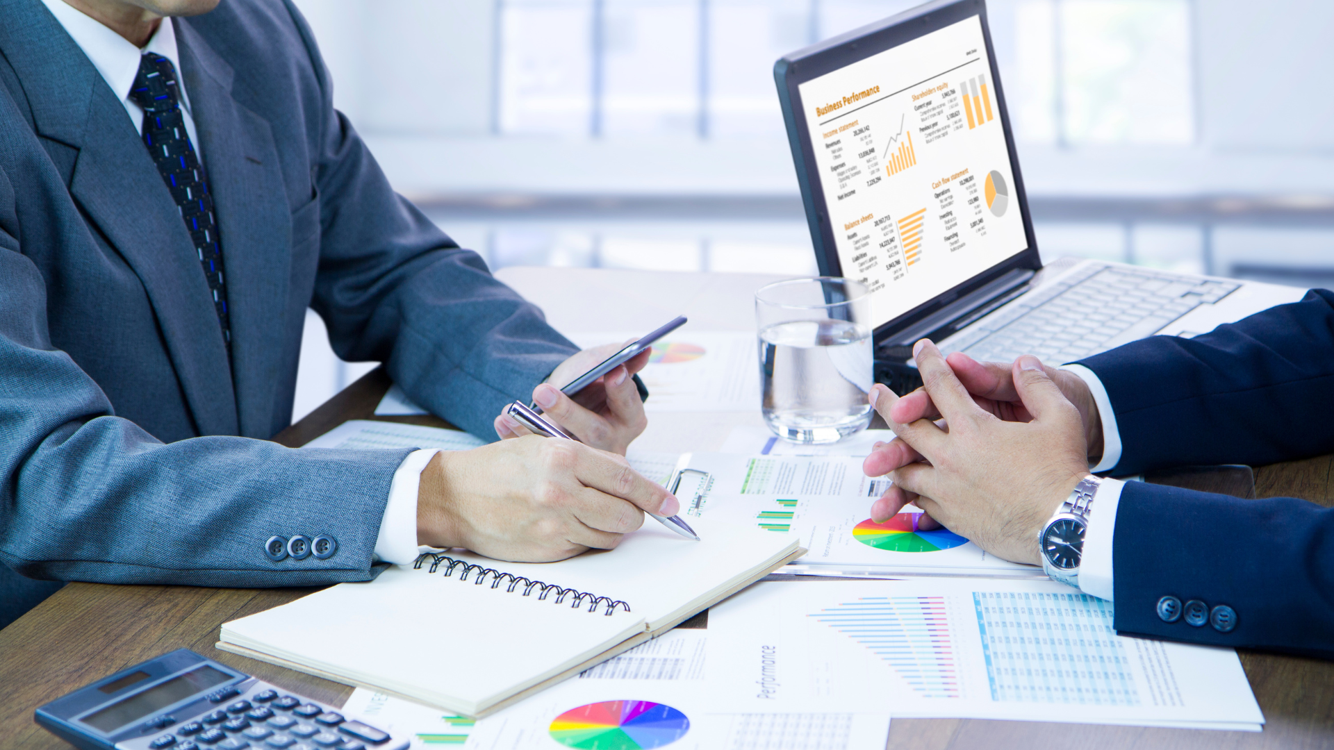 Two businessmen are sitting at a table with a laptop and a calculator.