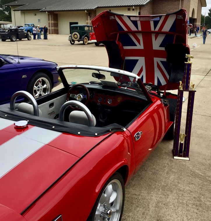 Red convertible with Union Jack flag display at an outdoor car show, with other cars and people in the background