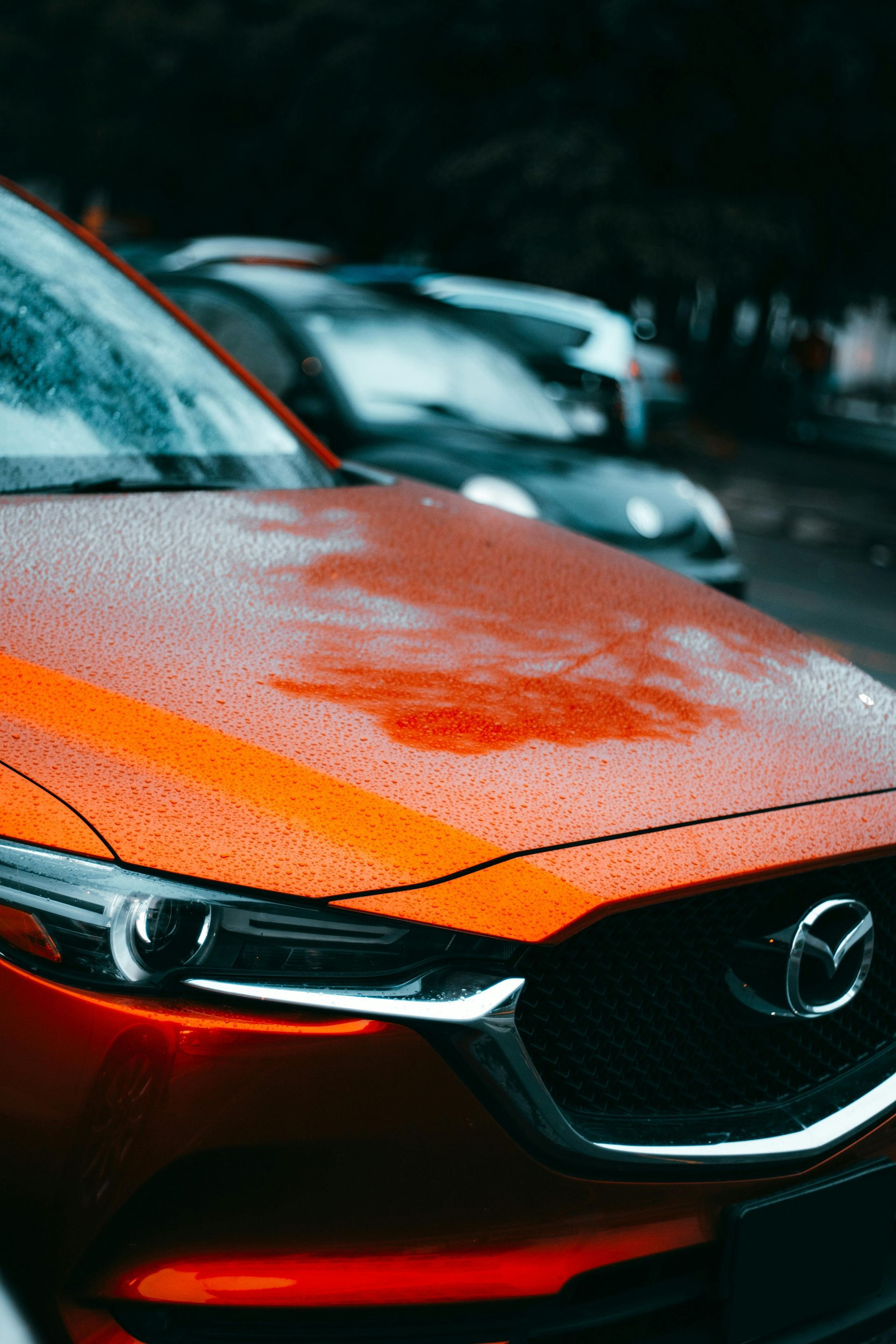 Close-up of a rain-speckled orange Mazda car front on a wet street with another car blurred behind.
