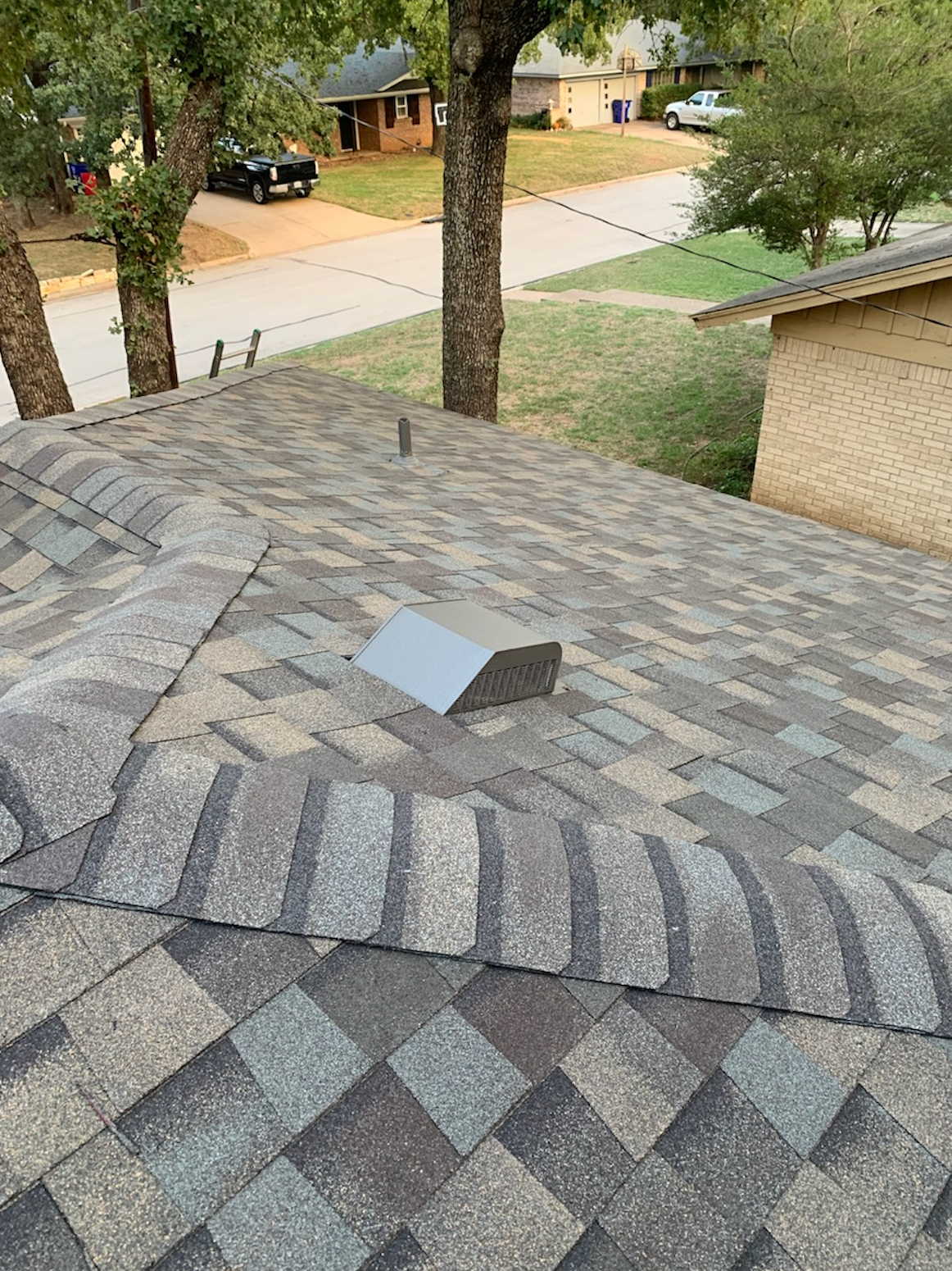 A roof with gray and brown shingles has a silver vent. Trees and houses are in the background.