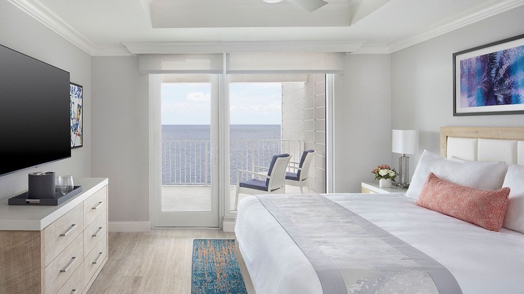 A hotel room with a king size bed , dresser , television and balcony overlooking the ocean.