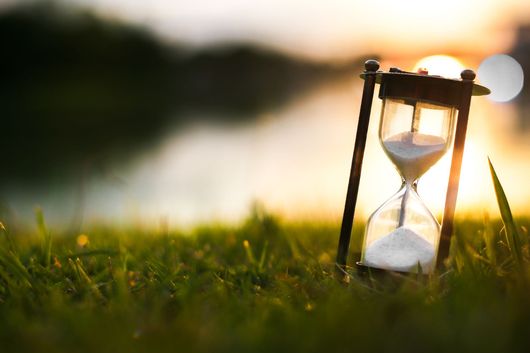 An hourglass rests in lush green grass at sunset, with a blurred lake and bright sun in the background.