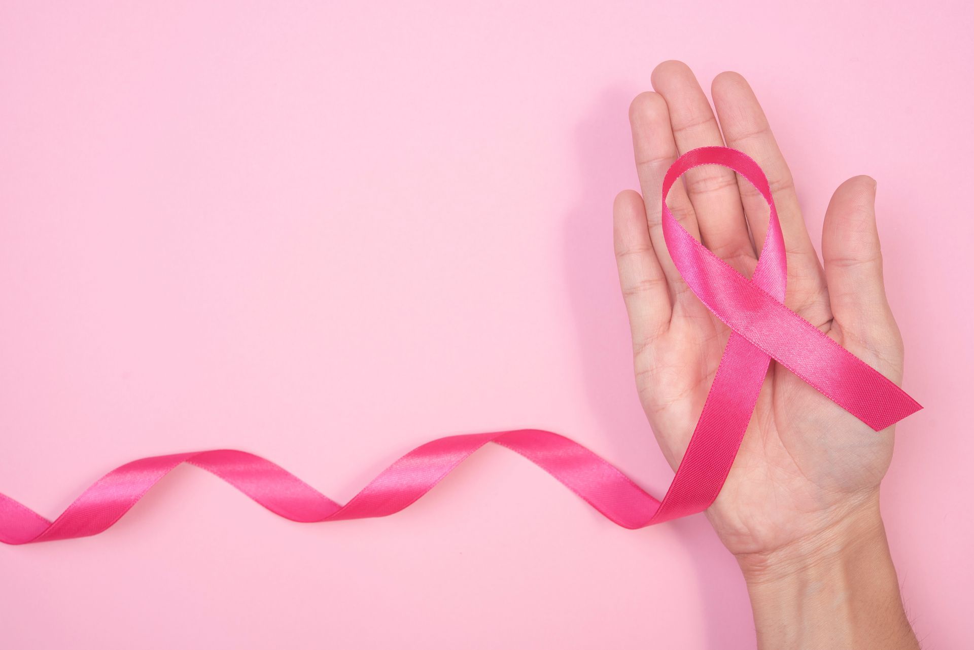 A pink awareness ribbon held in an open palm against a light pink background.