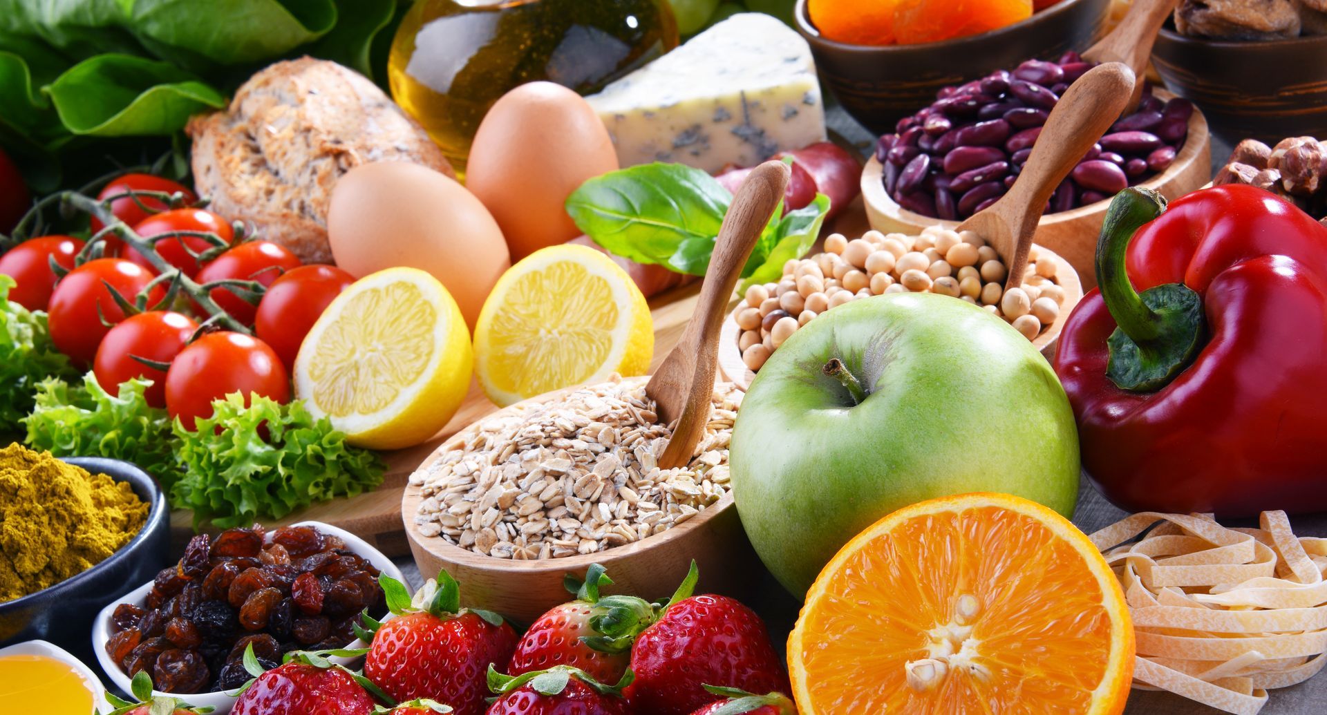 A colorful assortment of fresh produce including tomatoes, eggs, strawberries, an apple, orange, beans, and grains.
