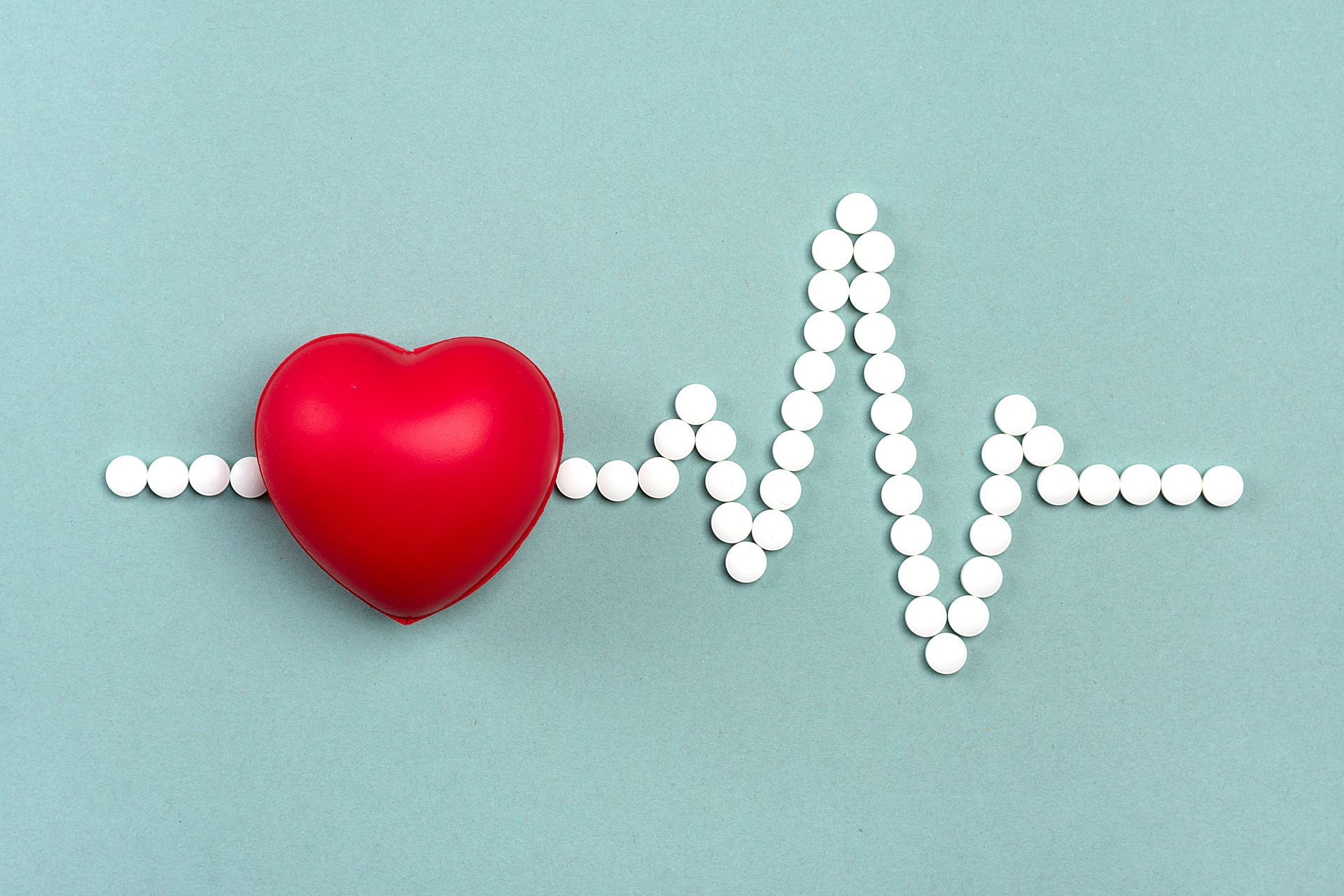 A red heart model sits on a line of white pills arranged to look like an EKG heart rate monitor on a blue background.