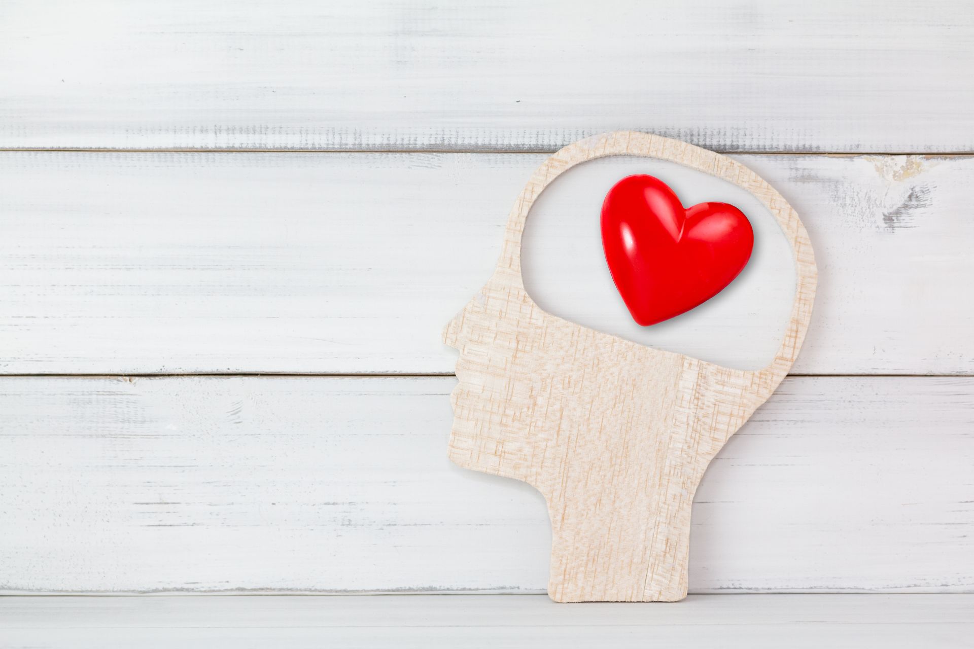 A wooden cutout of a human head profile on a white wooden background, with a red heart icon inside the brain area.