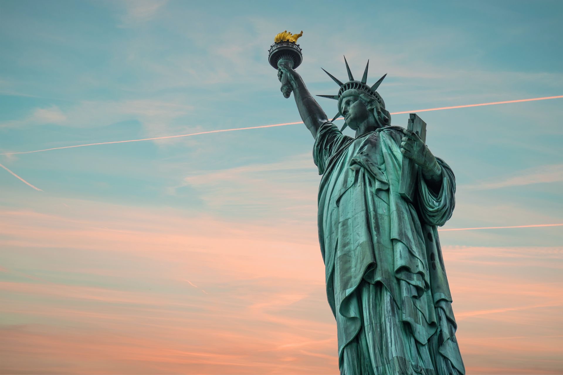 The Statue of Liberty holding a torch against a sunset sky with visible airplane vapor trails.