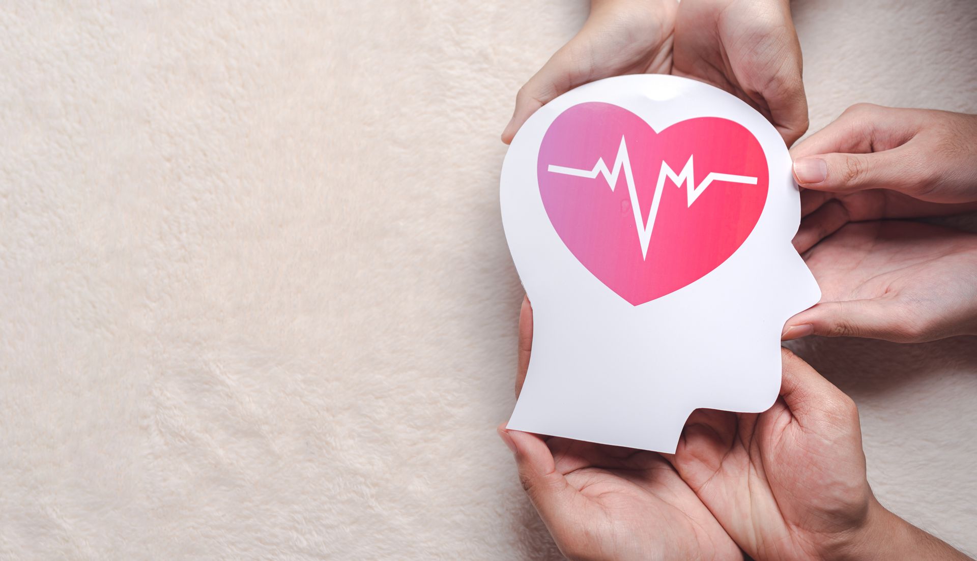 Two pairs of hands holding a paper cutout of a human head silhouette featuring a pink heart with an EKG line.