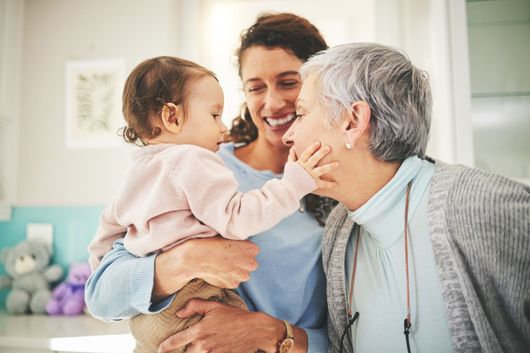 A smiling woman holding a baby who is reaching out to touch the face of another smiling woman.