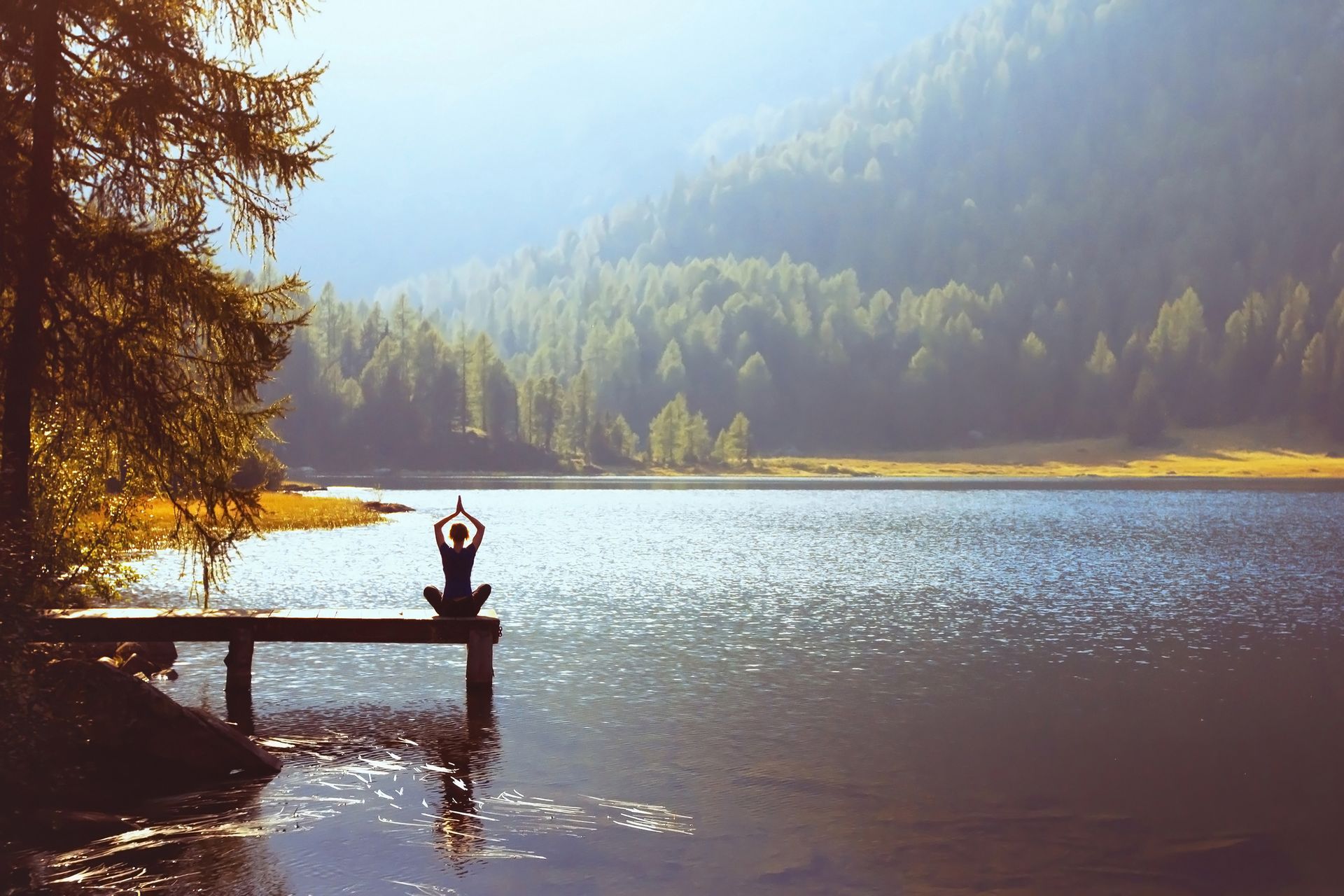 A person sits in a meditative pose on a wooden pier extending into a calm lake, backed by a lush forest and mountains.