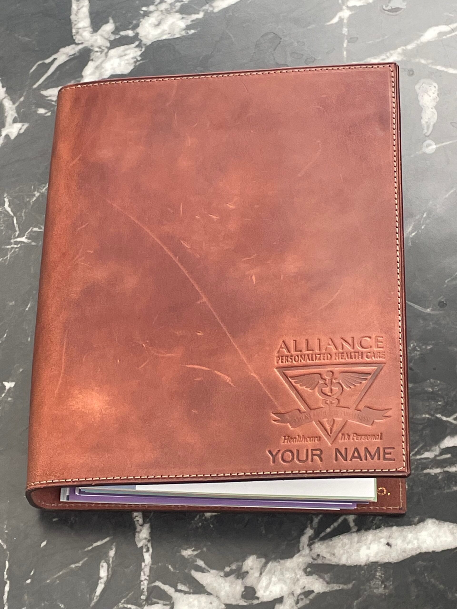 Brown leather-bound planner on a dark stone surface, with an embossed logo and the words “Your Name.”