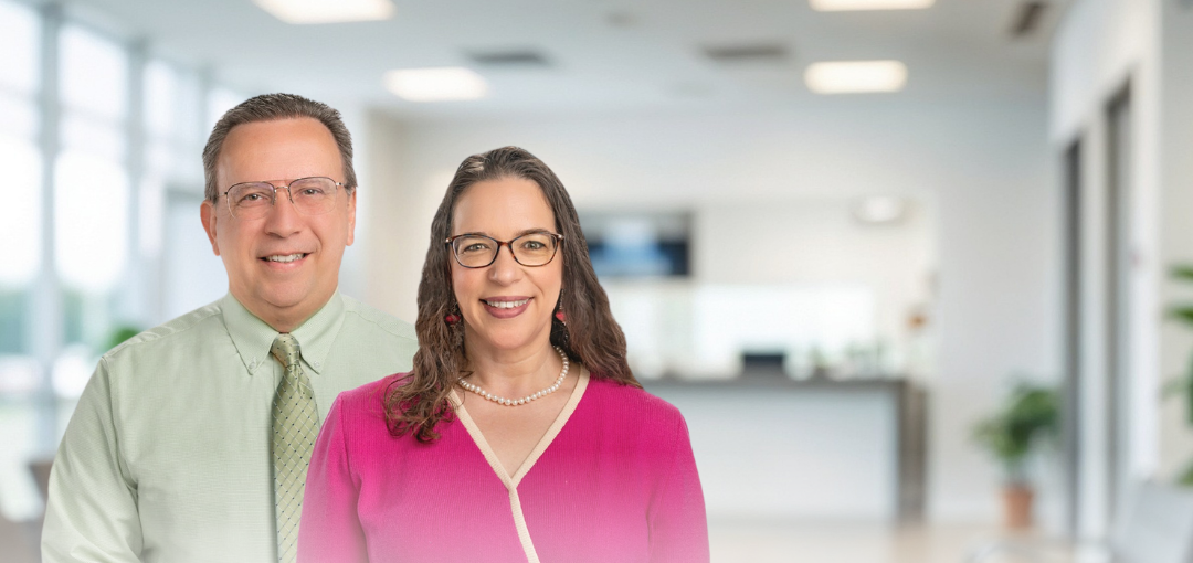A man in a light green shirt and a woman in a pink top stand together in a blurred, bright, modern office lobby.