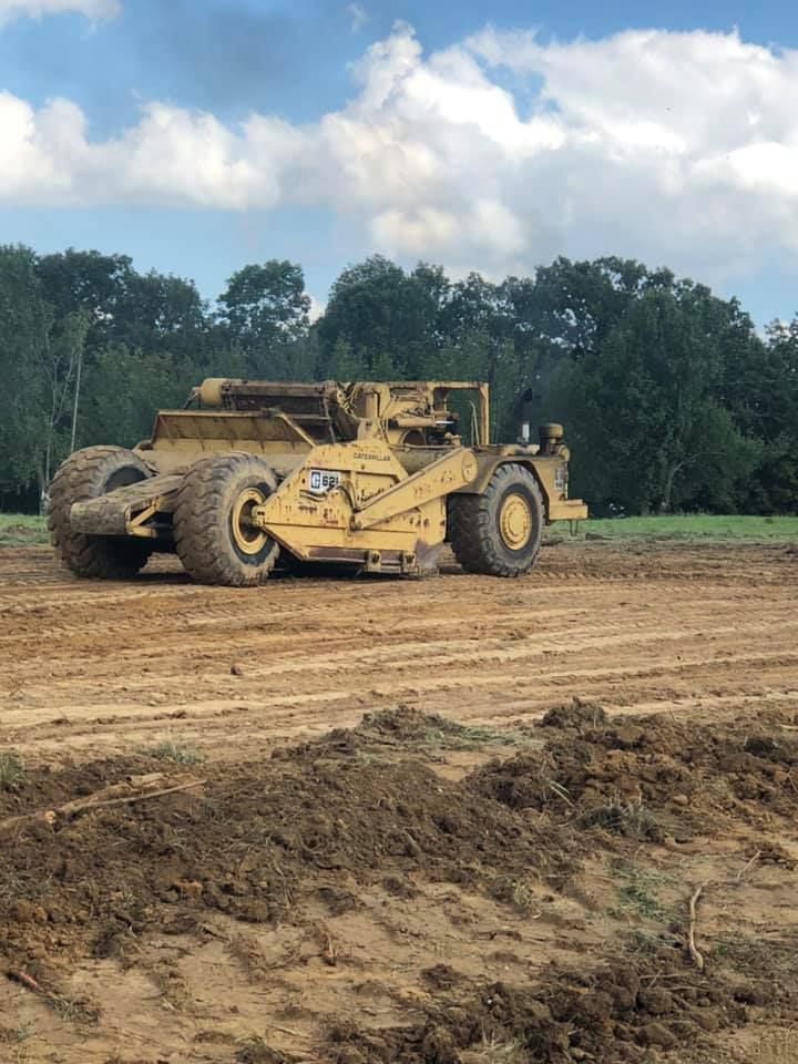 A large yellow earth-moving scraper machine operating on a dirt field under a cloudy sky.