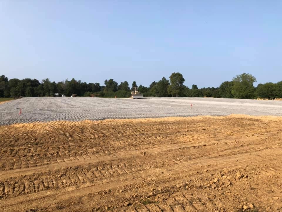A wide, dirt-covered construction site with a large, gravel-filled area in the center, bordered by distant trees.