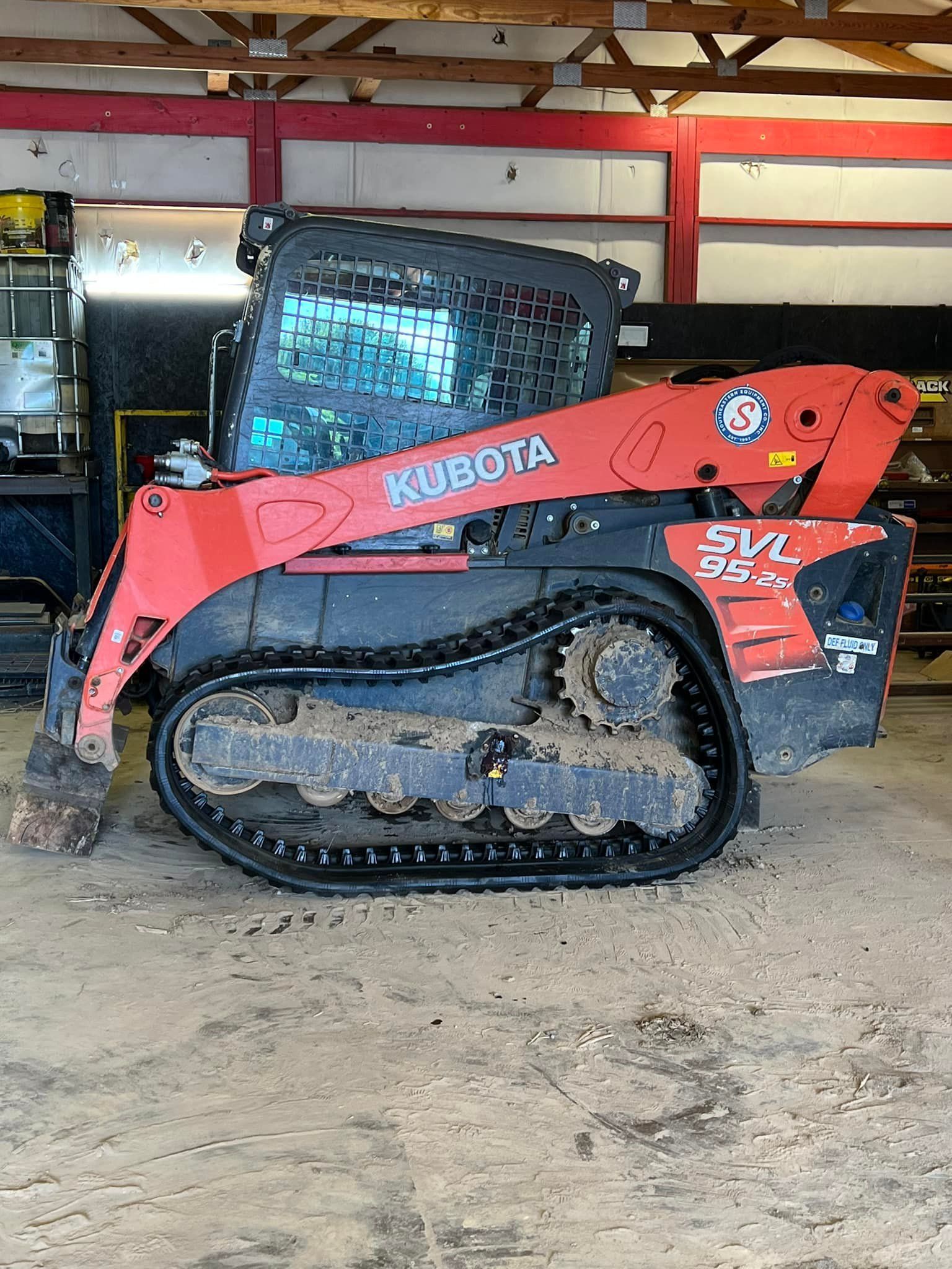 A red Kubota SVL95 compact track loader parked inside a building with a corrugated metal ceiling and red beams.
