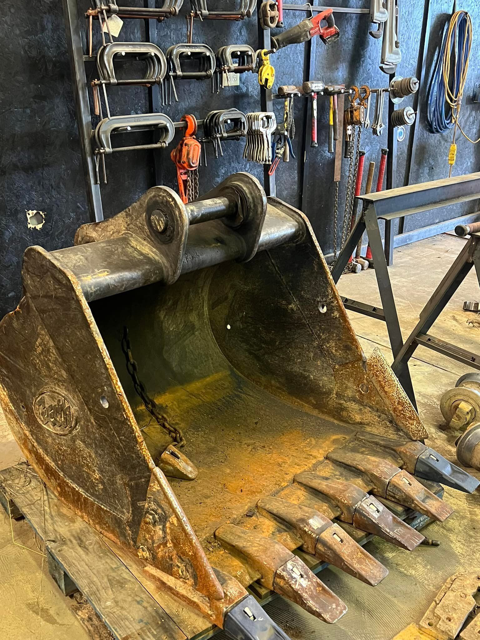 A rusted excavator bucket with several metal teeth sits on a wooden pallet in a workshop with tools hanging on a wall.
