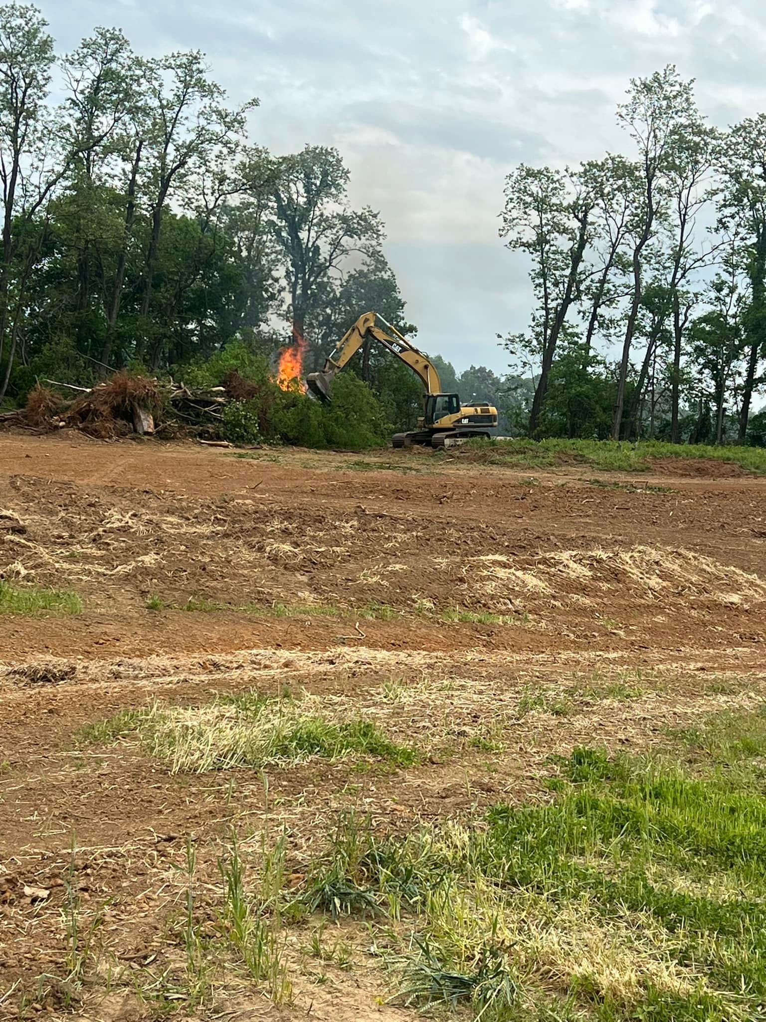 A yellow excavator works in a cleared, dirt-covered field near trees while a pile of brush burns in the background.