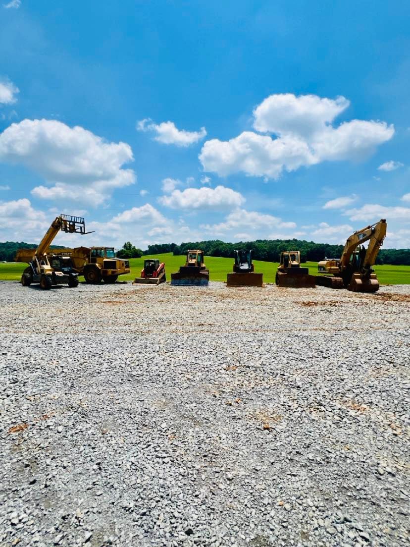 A line of heavy construction equipment parked on a gravel lot under a bright, cloudy blue sky.